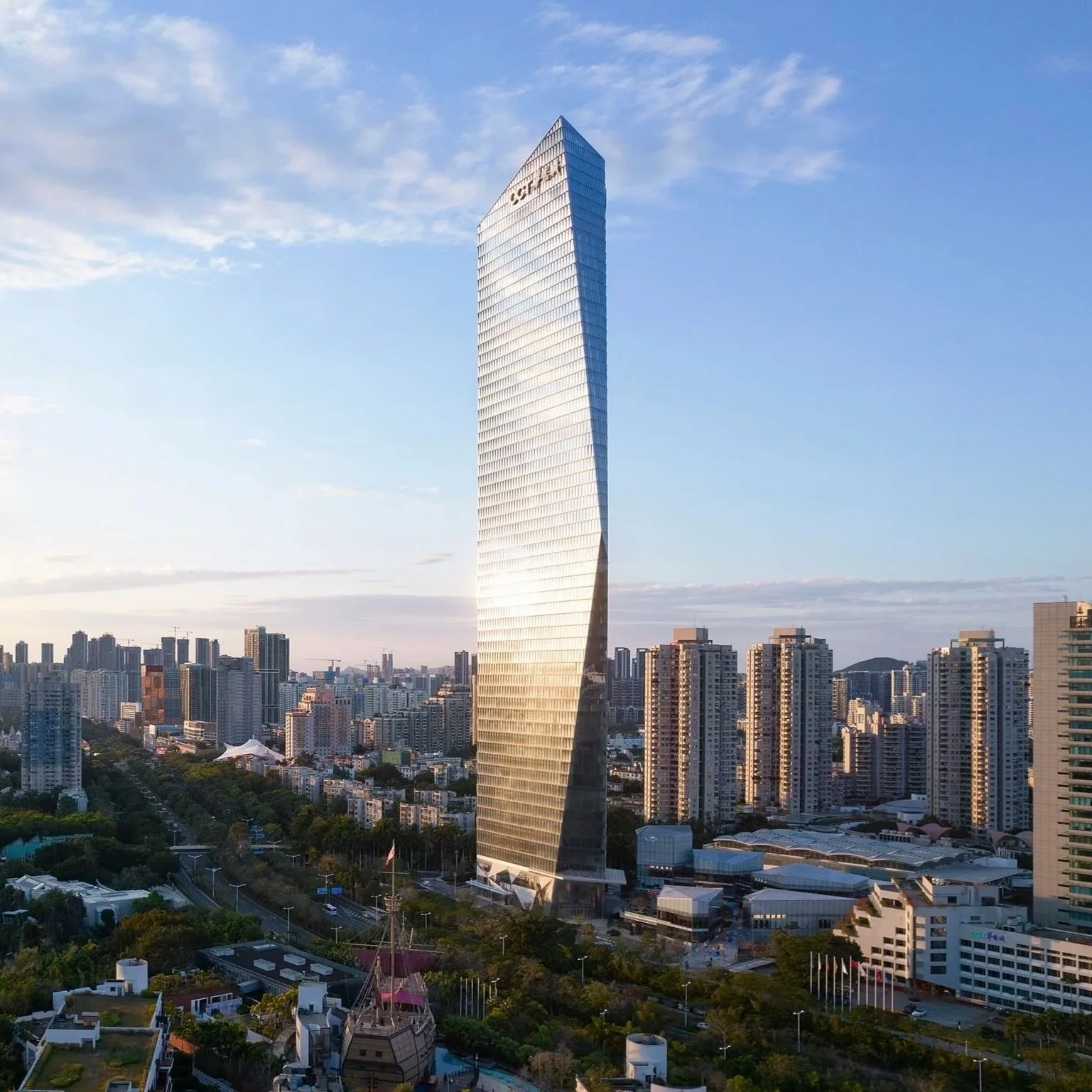 Tall modern skyscraper with a reflective glass facade against a blue sky, surrounded by numerous high-rise buildings in an urban cityscape.