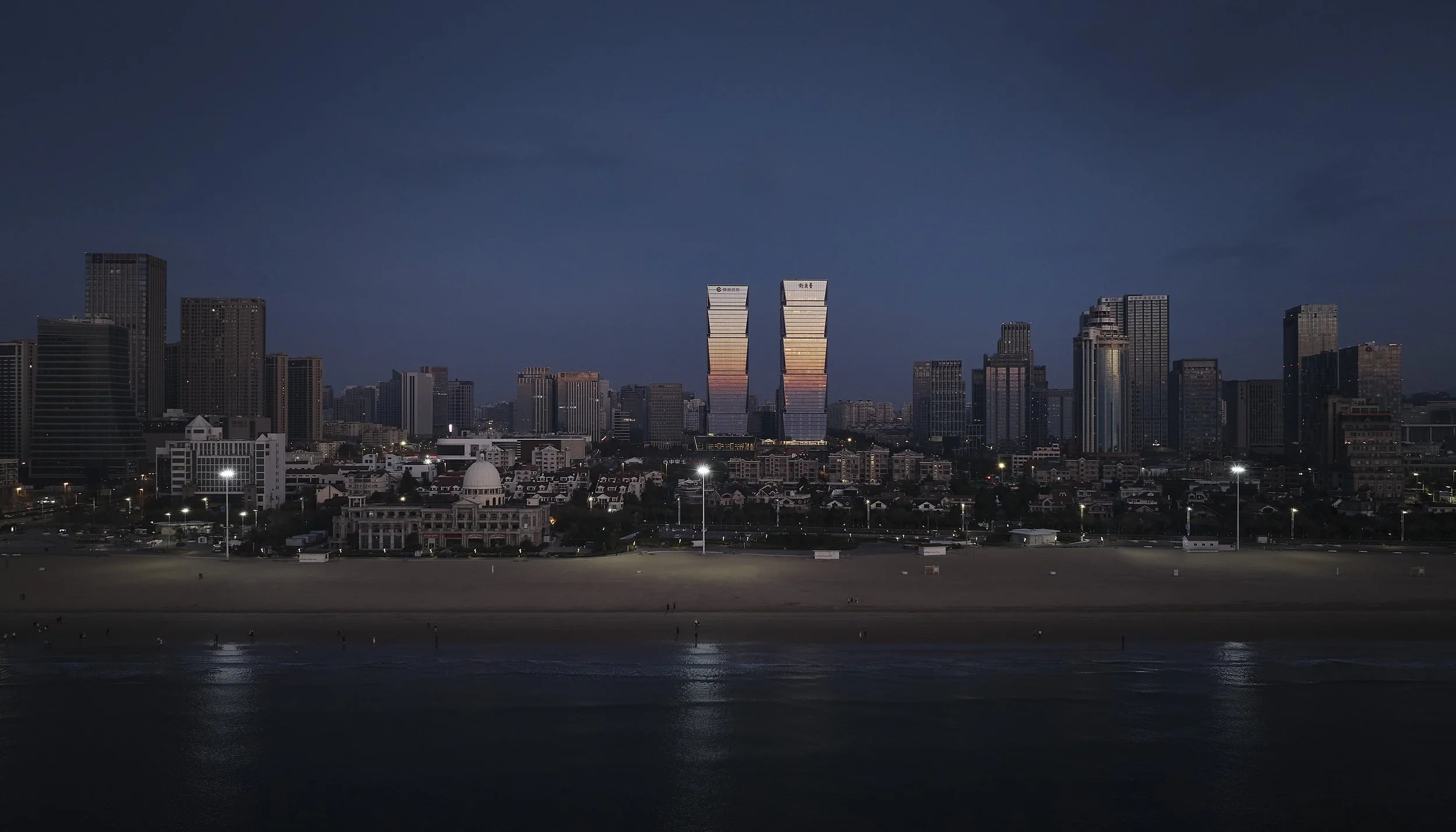 City skyline at dusk with high-rise buildings reflecting in the water, and a beach in the foreground.