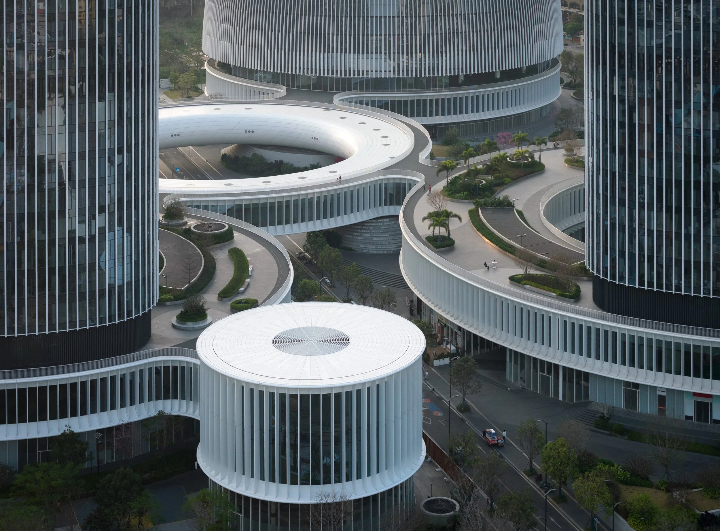 Modern office building with curved white levels and vertical slats, surrounded by landscaped greenery and walkways.