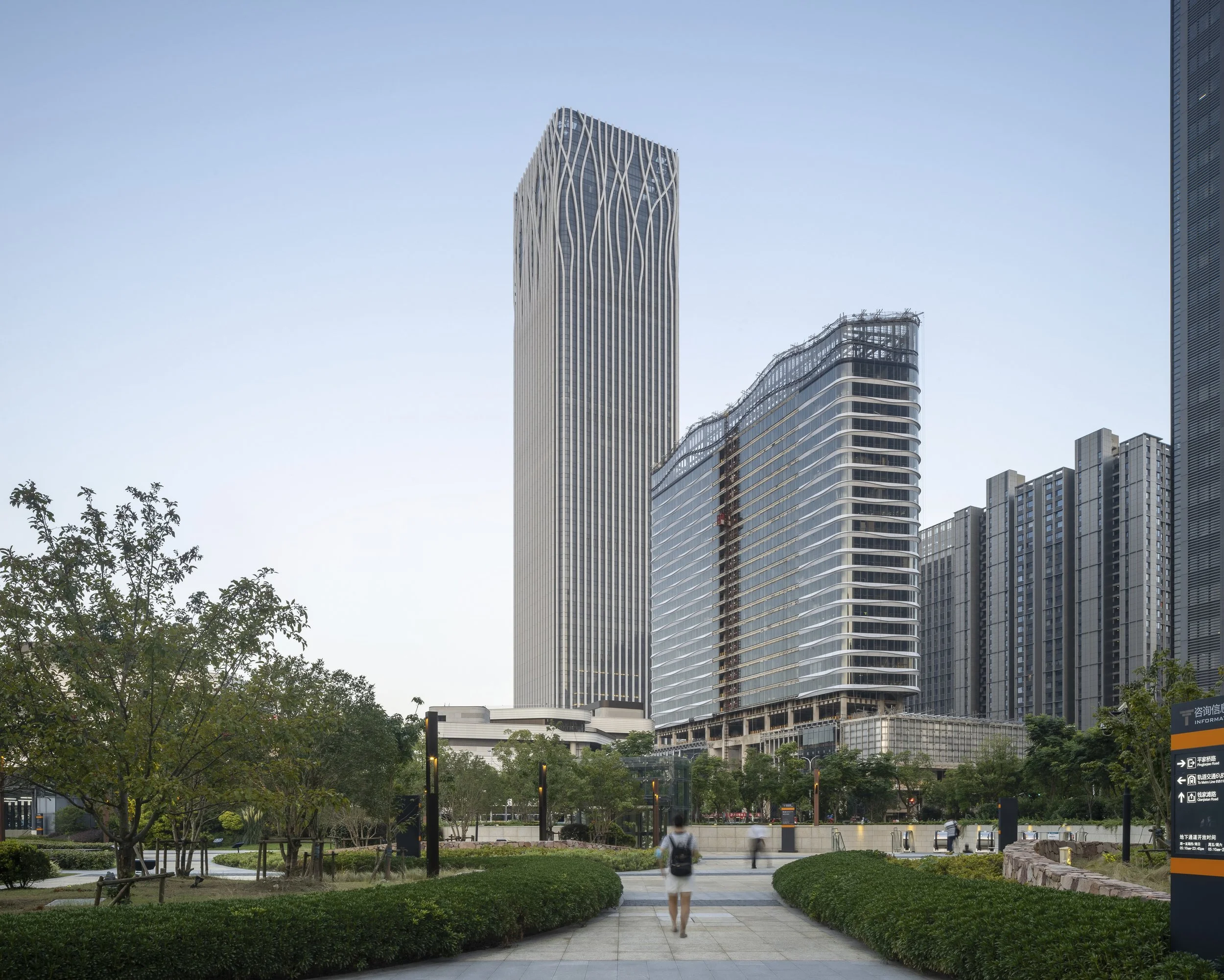 Urban park with trees and pathways in front of modern skyscrapers, including one tall building with a wave design on its facade, under a clear sky.