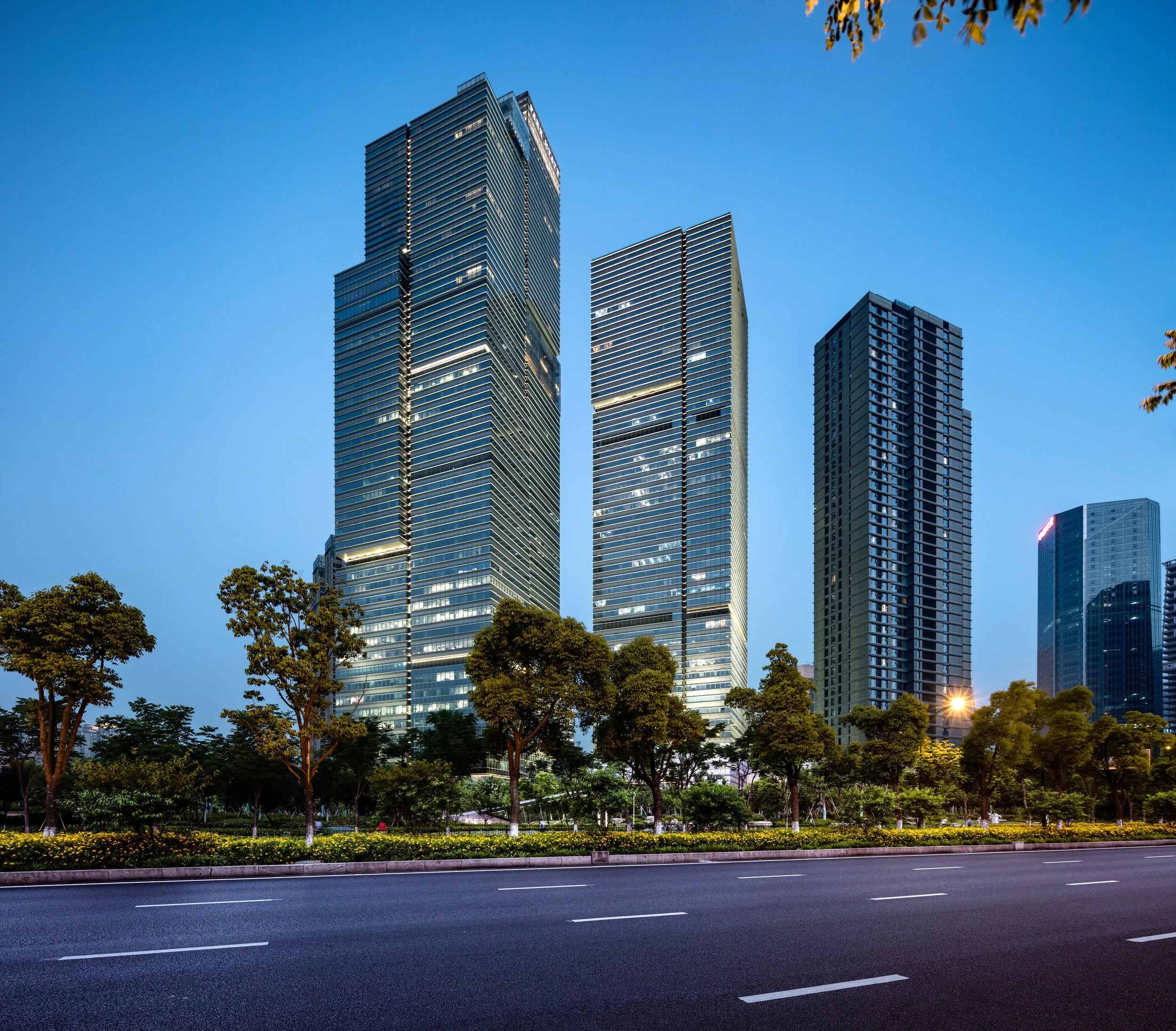 Photo of modern skyscrapers against a blue sky, with trees and a road in the foreground.