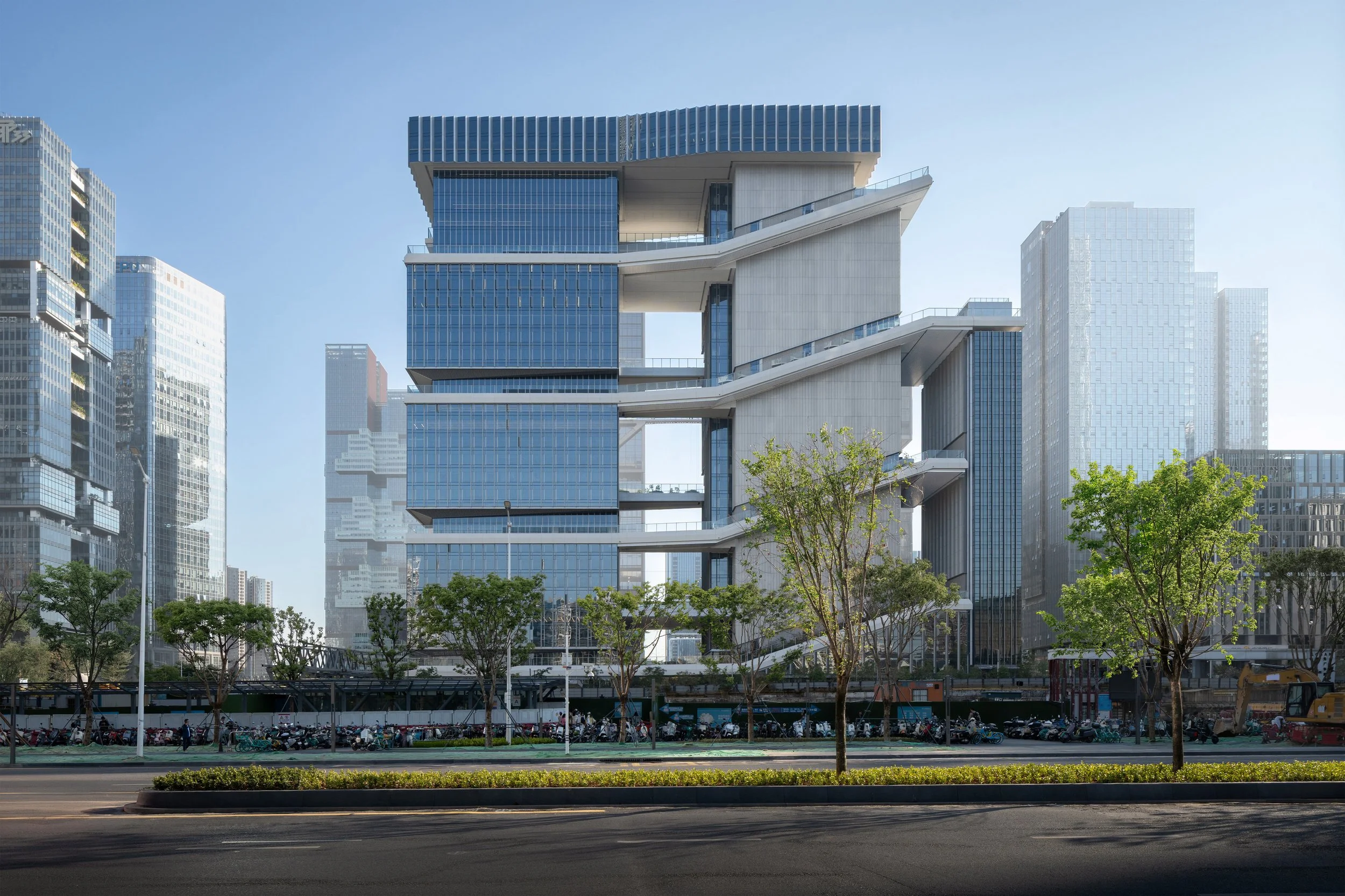 Modern high-rise building with unique architectural design in a cityscape, surrounded by other tall buildings, with trees and a parking area in the foreground.