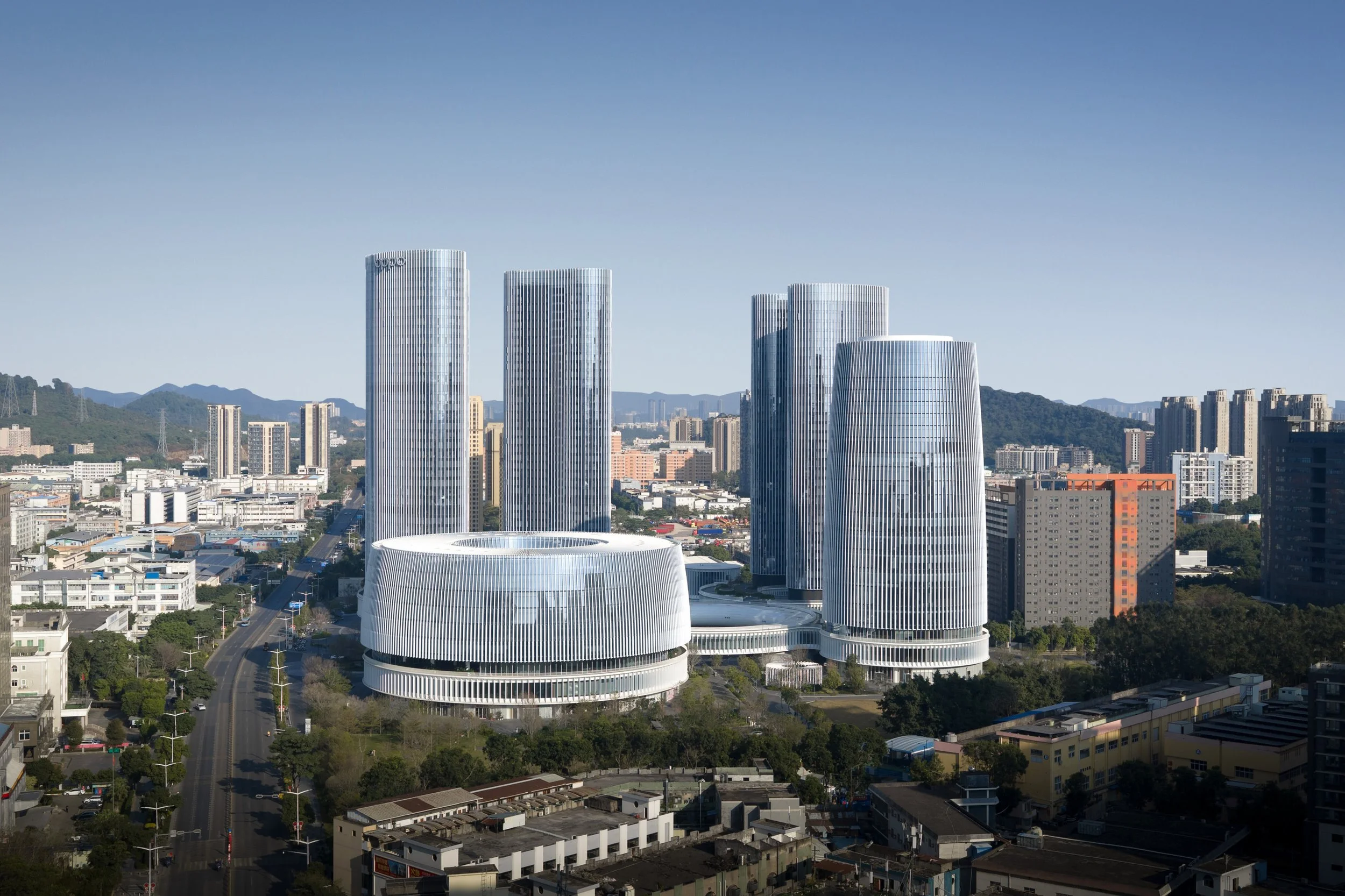 A city skyline with modern glass skyscrapers against a backdrop of green hills and mountains, under a clear blue sky.