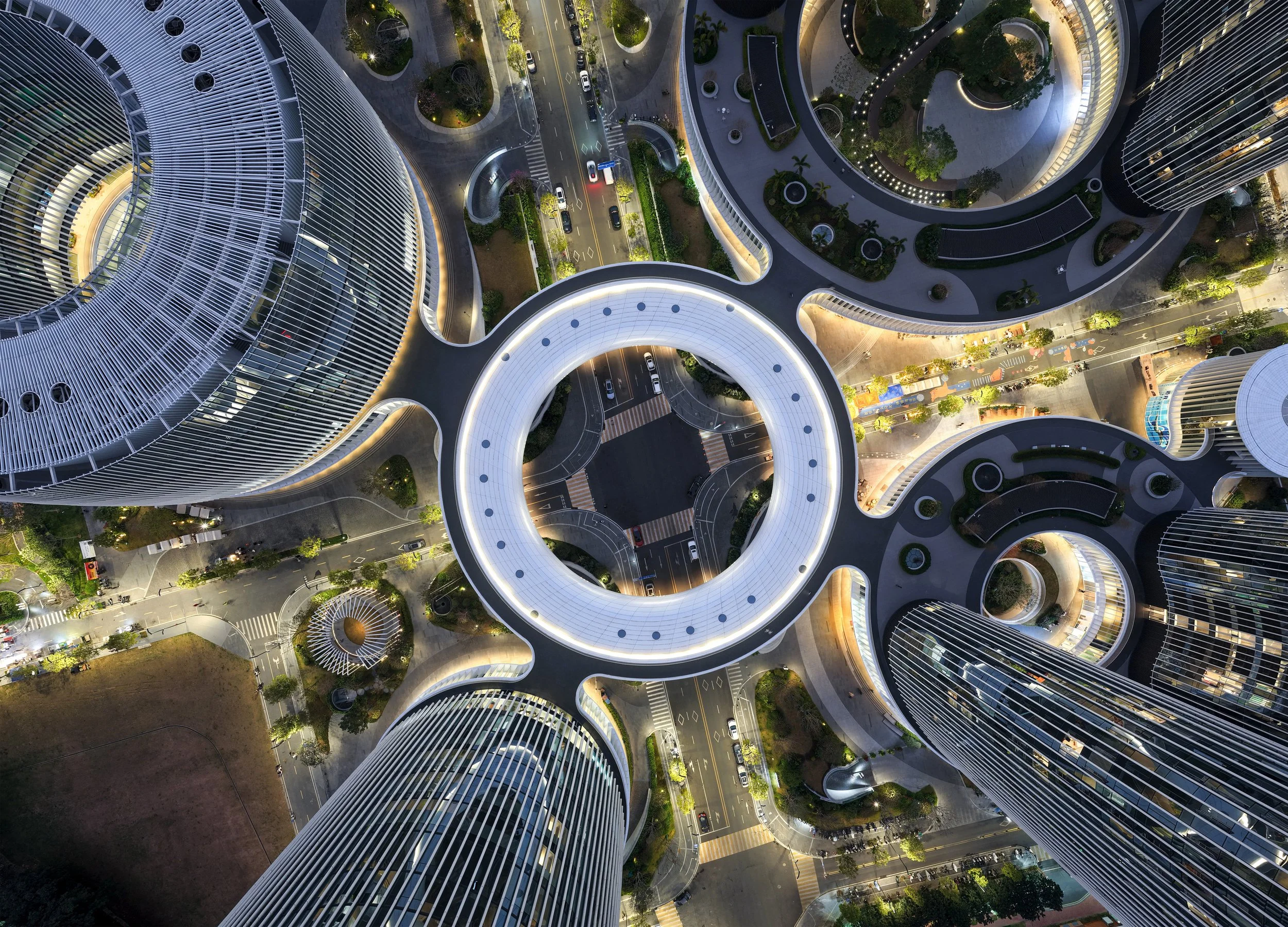 Aerial view of modern high-rise buildings with circular and curved designs, illuminated by lights at dusk, with streets, trees, and vehicles visible below.