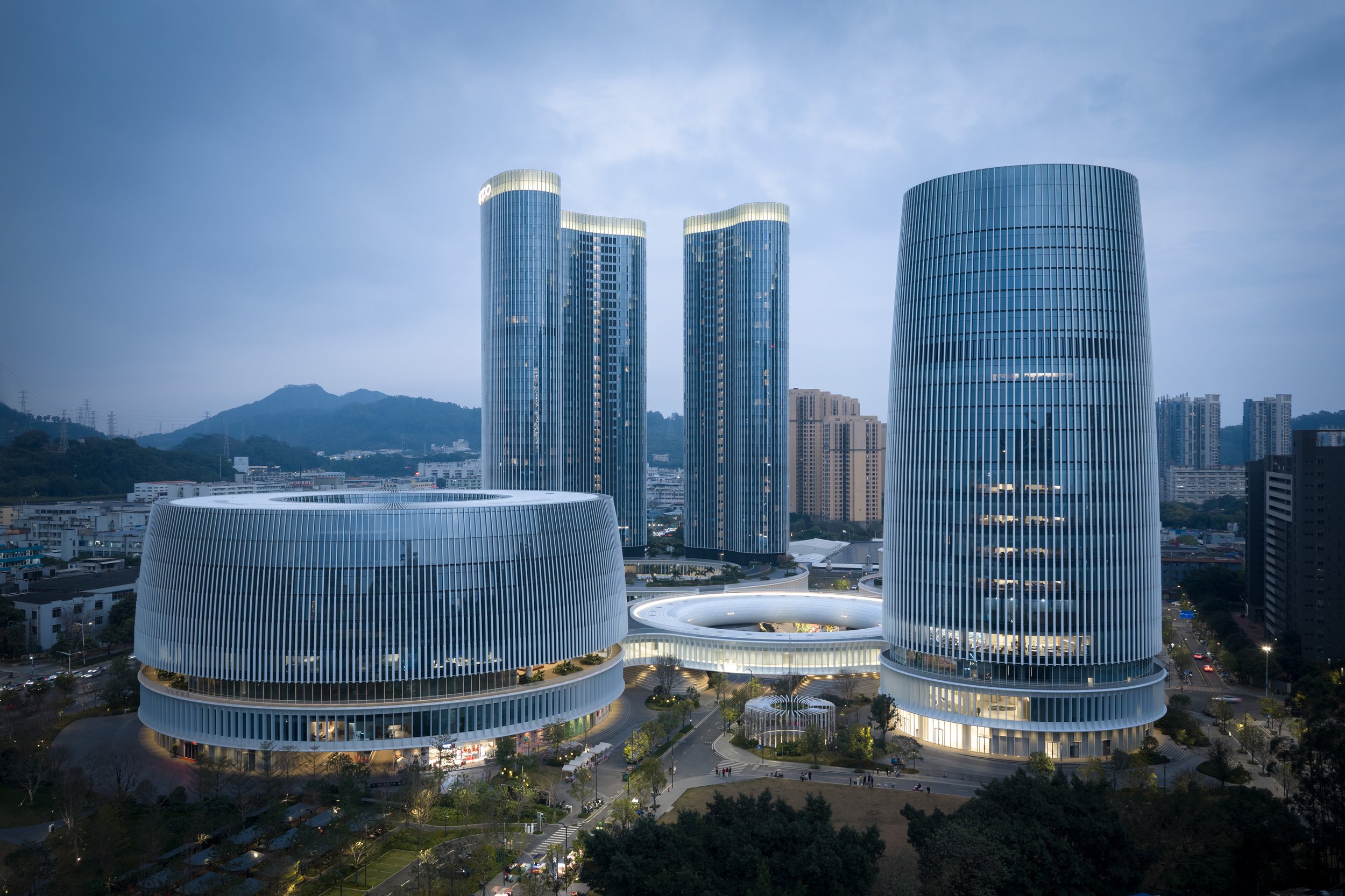 Cityscape featuring modern, tall glass skyscrapers with rounded and curved designs, set against a backdrop of mountains and a cloudy sky at dusk.