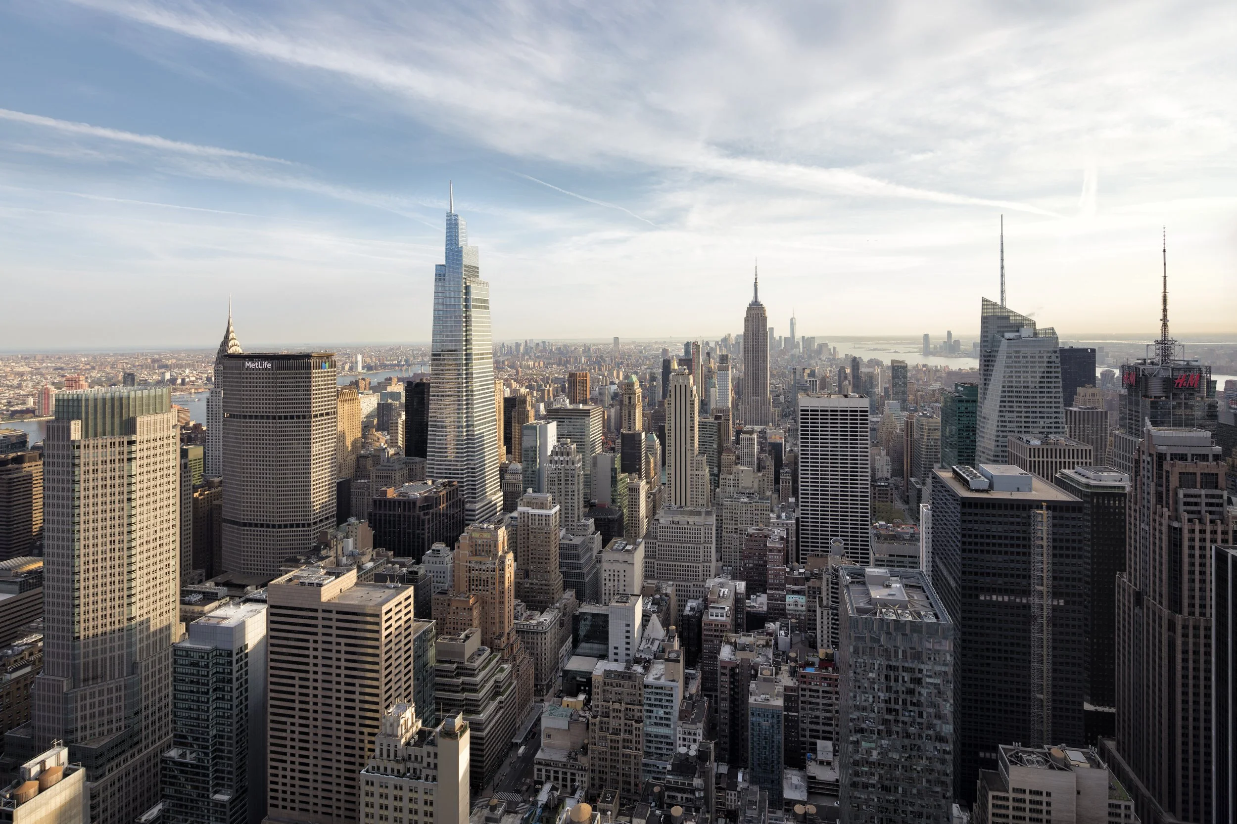 A city skyline with numerous tall skyscrapers, including the Empire State Building, under a partly cloudy sky in New York City.