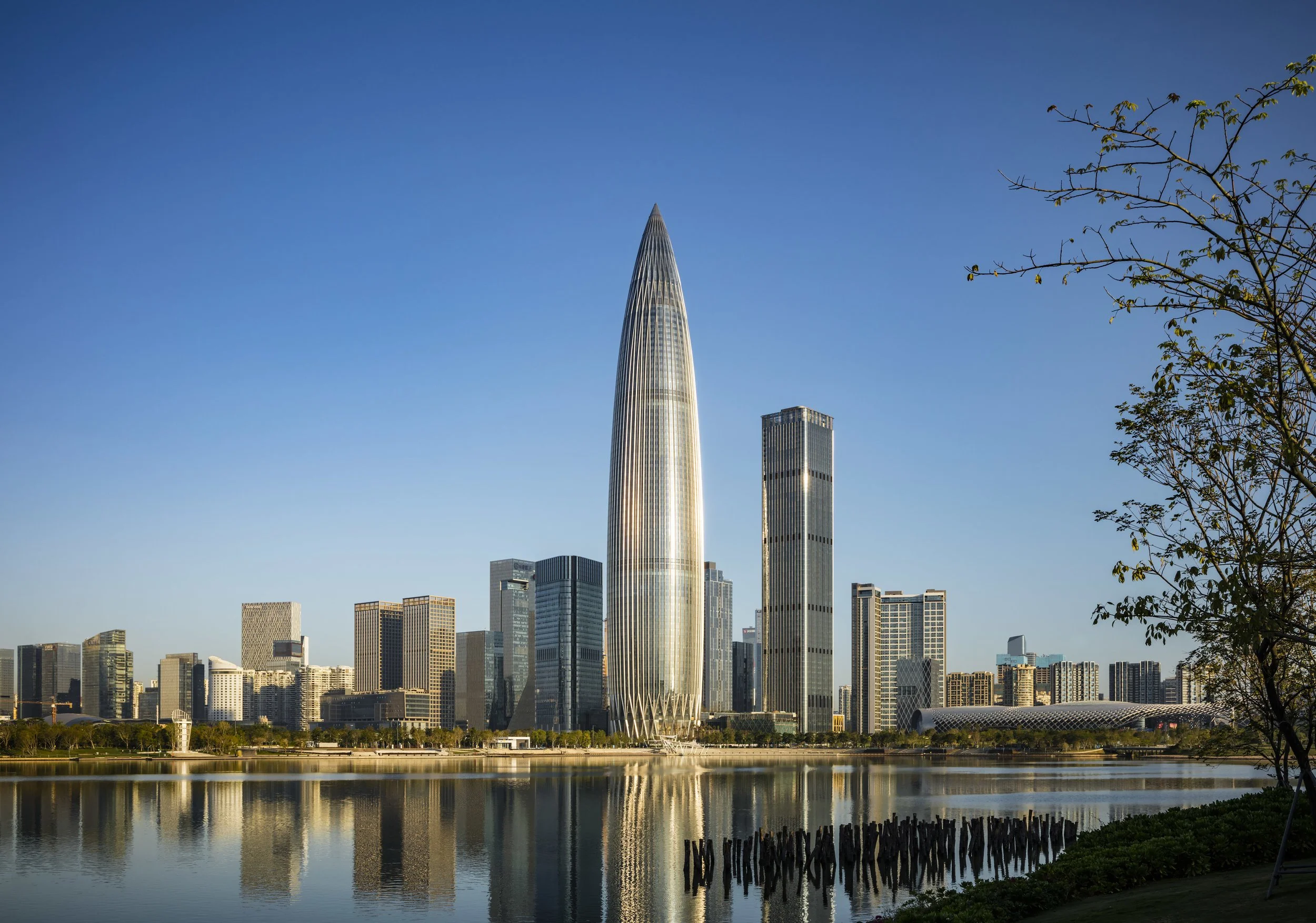 City skyline featuring tall, modern skyscrapers including a distinctive, pointed tower, reflected in a calm body of water with trees and greenery in the foreground, under a clear blue sky.