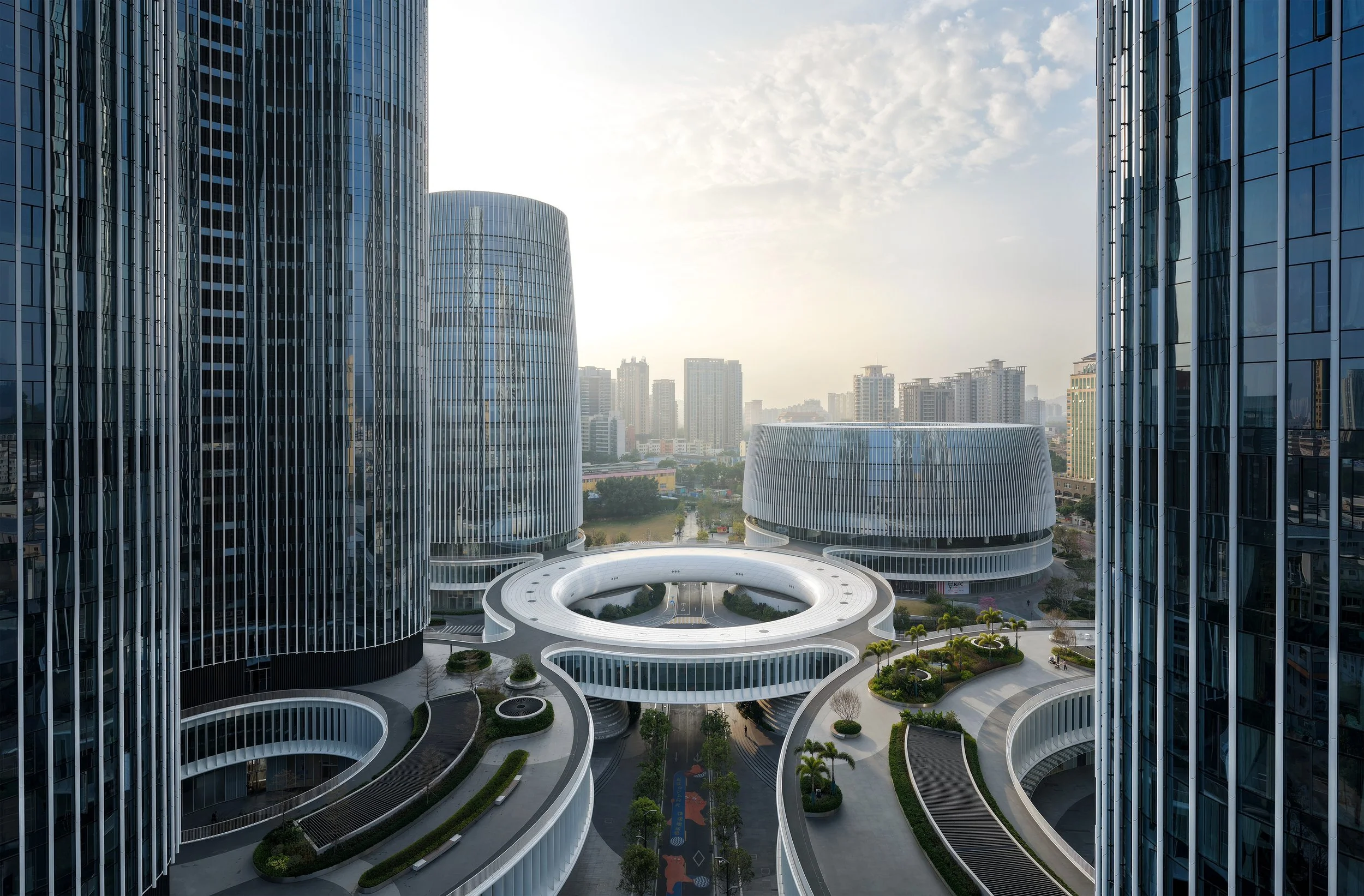 Modern cityscape with tall glass buildings, curved architectural structures, and a landscaped courtyard with trees and walking paths.
