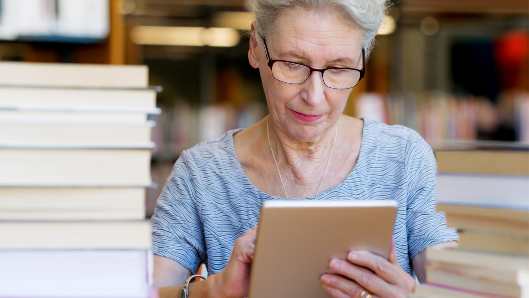 An elderly woman with glasses reading a tablet in a library, surrounded by stacks of books.