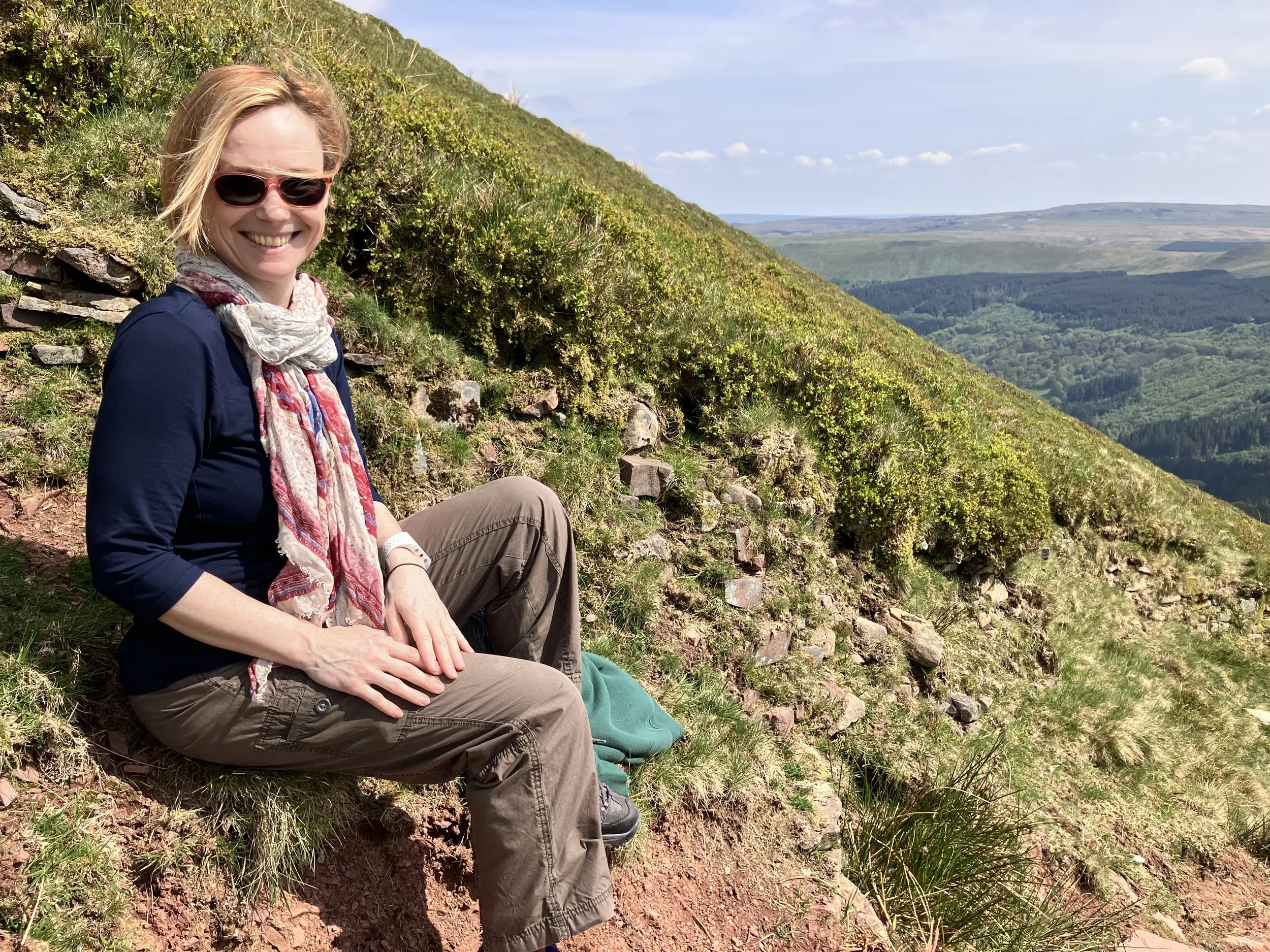 A woman wearing sunglasses, a scarf, and outdoor clothing sitting on a hillside with lush green vegetation, overlooking a valley with distant hills and a blue sky with scattered clouds.