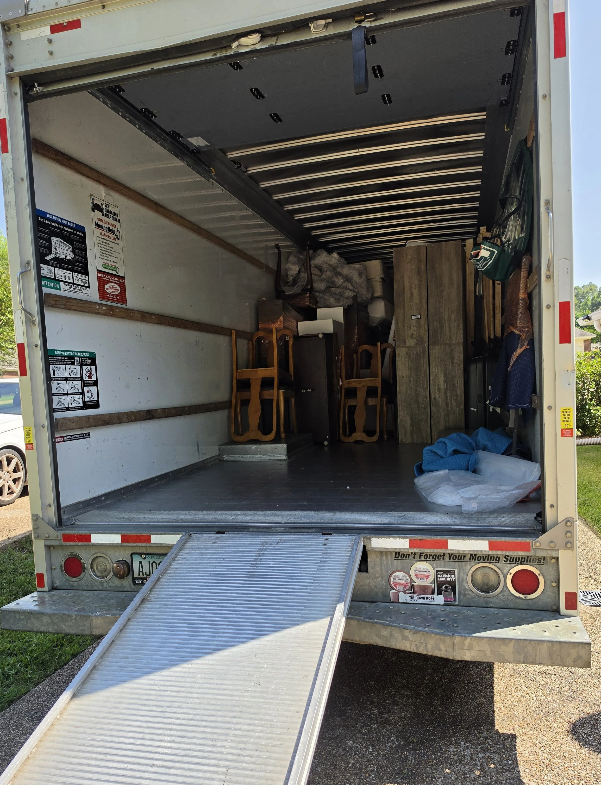 Open moving truck with a ramp extended, showing furniture and items inside, including chairs, cabinets, and storage bags.