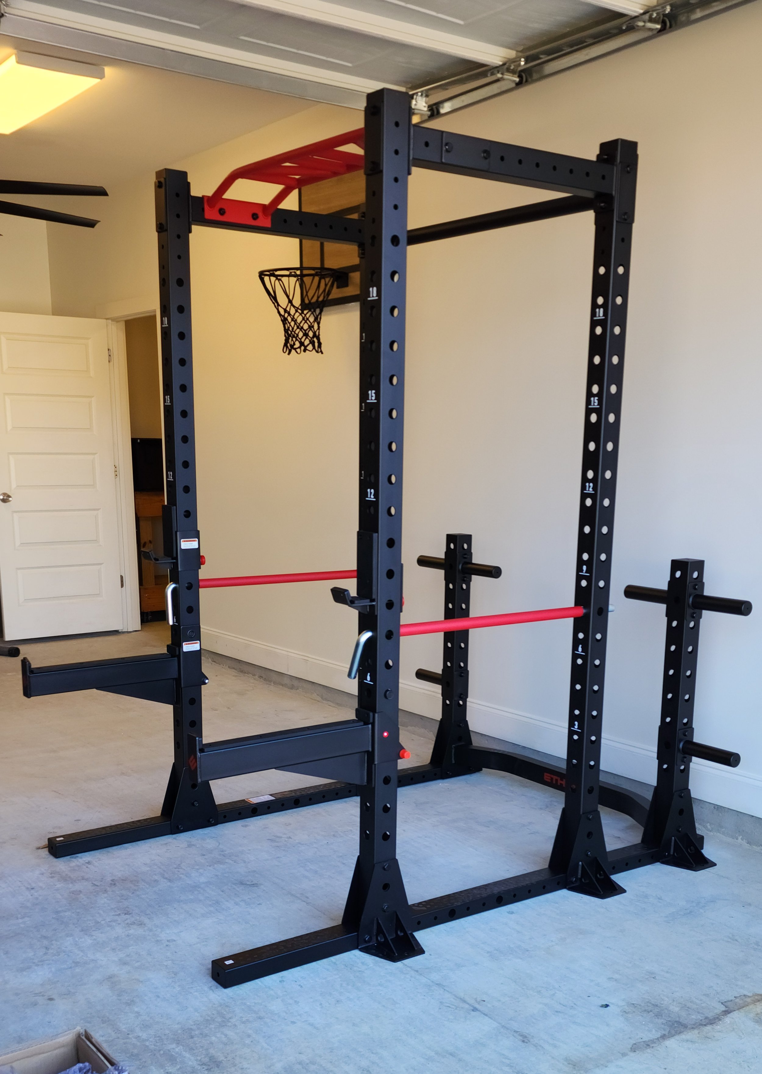 A home gym setup with a black and red power rack, a small basketball hoop attached to the rack, and weight bars on the sides. The room has a concrete floor, a closed door, and ceiling-mounted garage door tracks.
