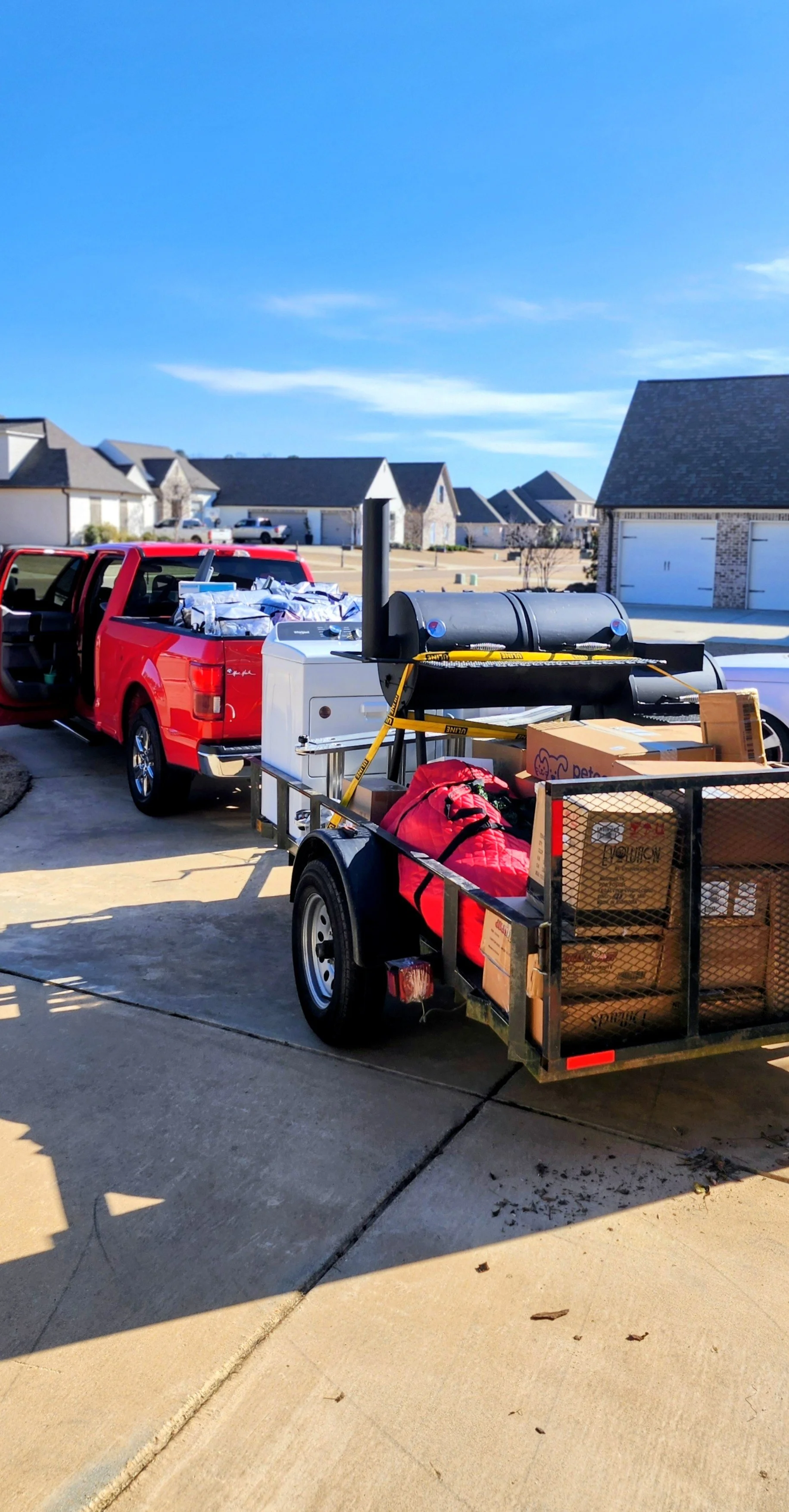 A red pickup truck with its tailgate down, attached to a cargo trailer filled with boxes, bags, and equipment parked on a driveway in front of suburban houses under a blue sky.
