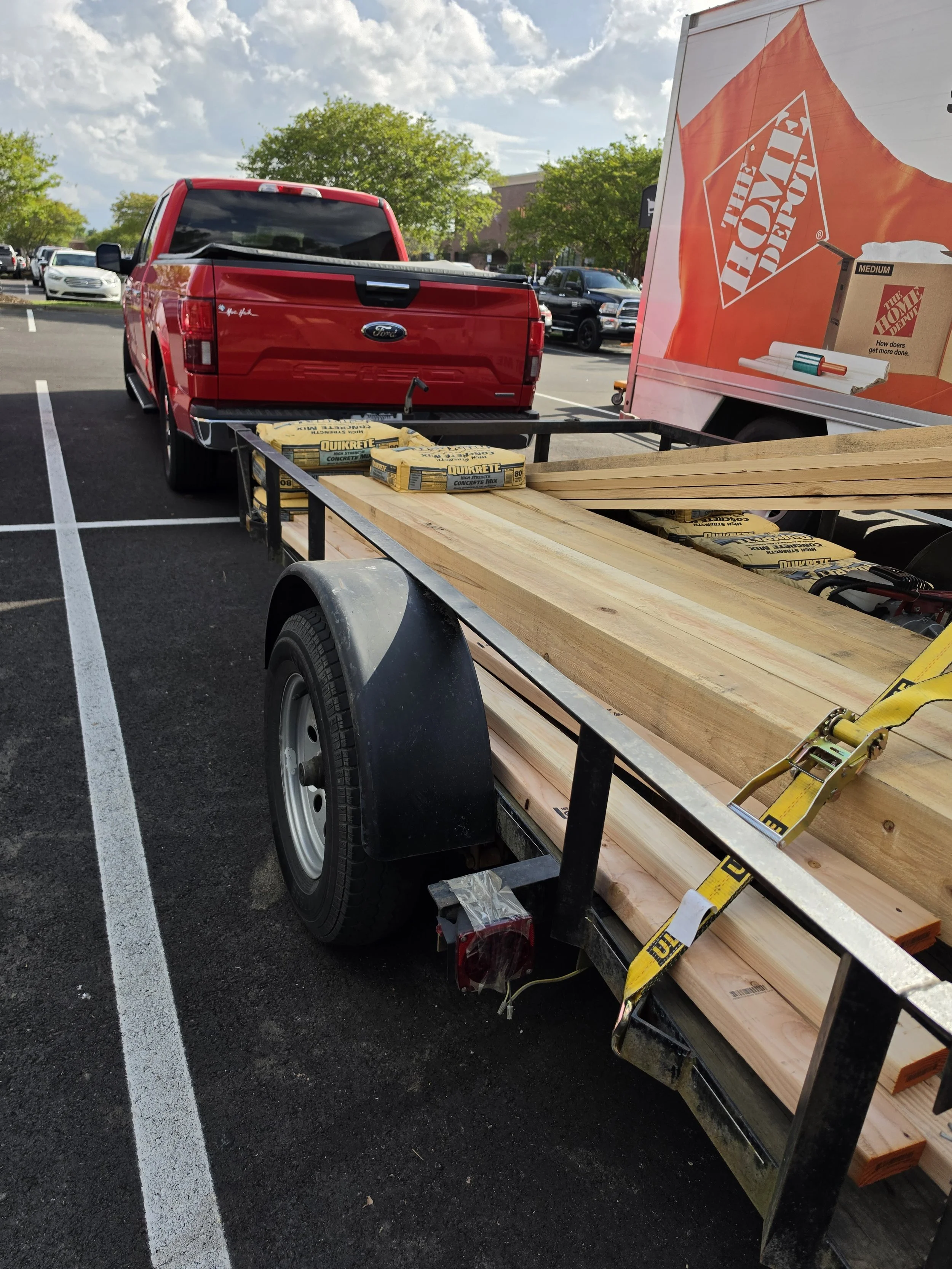 A red Ford truck with a trailer attached, loaded with pieces of lumber and bags of concrete mix, parked in a parking lot next to a Home Depot store.