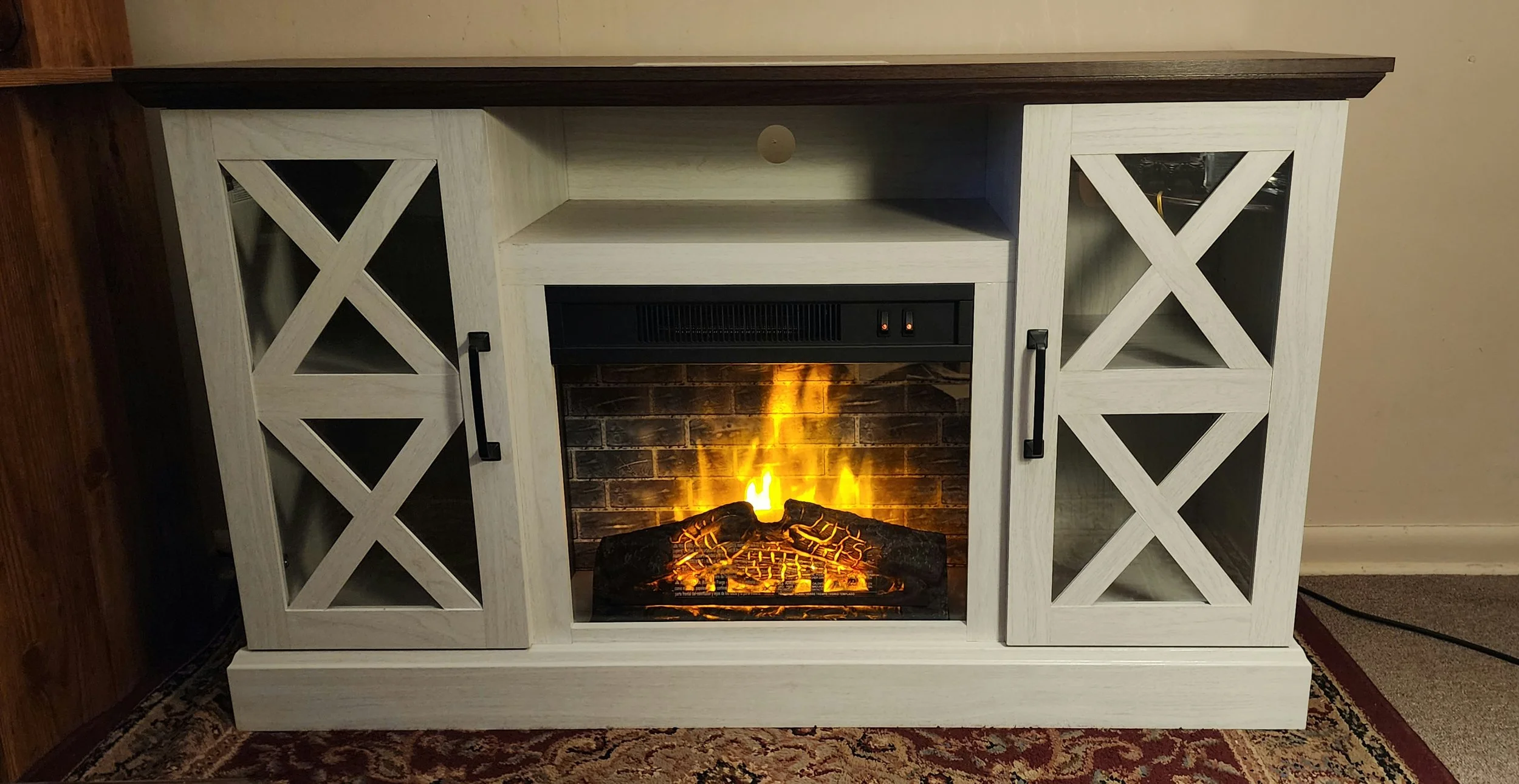 White, wooden electric fireplace with glass doors on either side, a black control panel in the middle, a faux brick interior with a glowing fire, on a patterned carpet in a room with beige walls.