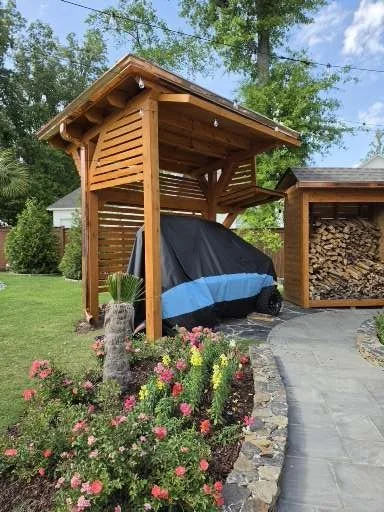 Wooden shed with a roof, housing a covered motorcycle in a backyard with flower bed and stone pathway