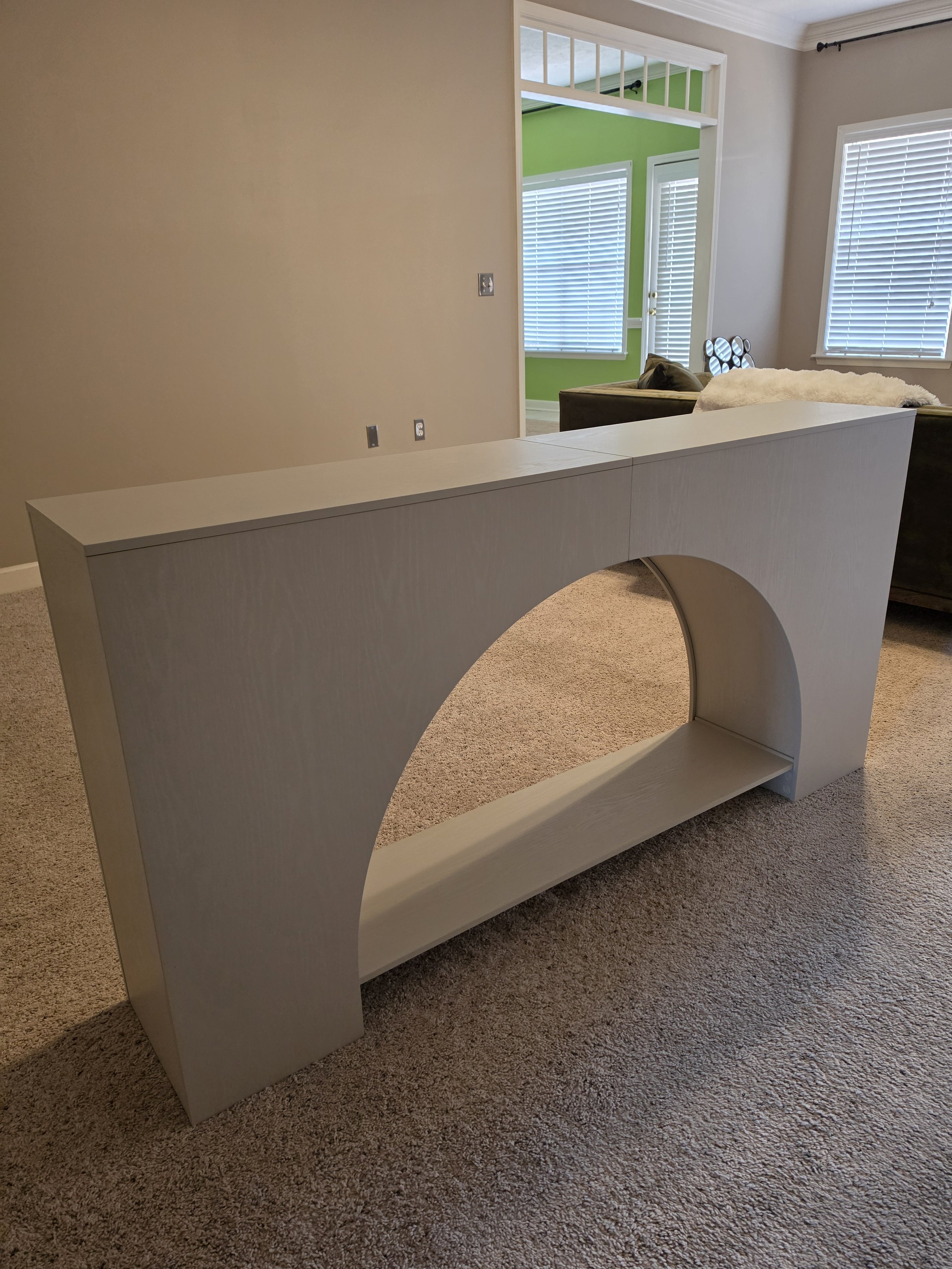 White modern curved bookshelf in beige carpeted living room with beige walls, adjacent to a sitting area with green walls, white blinds, and a door with blinds.