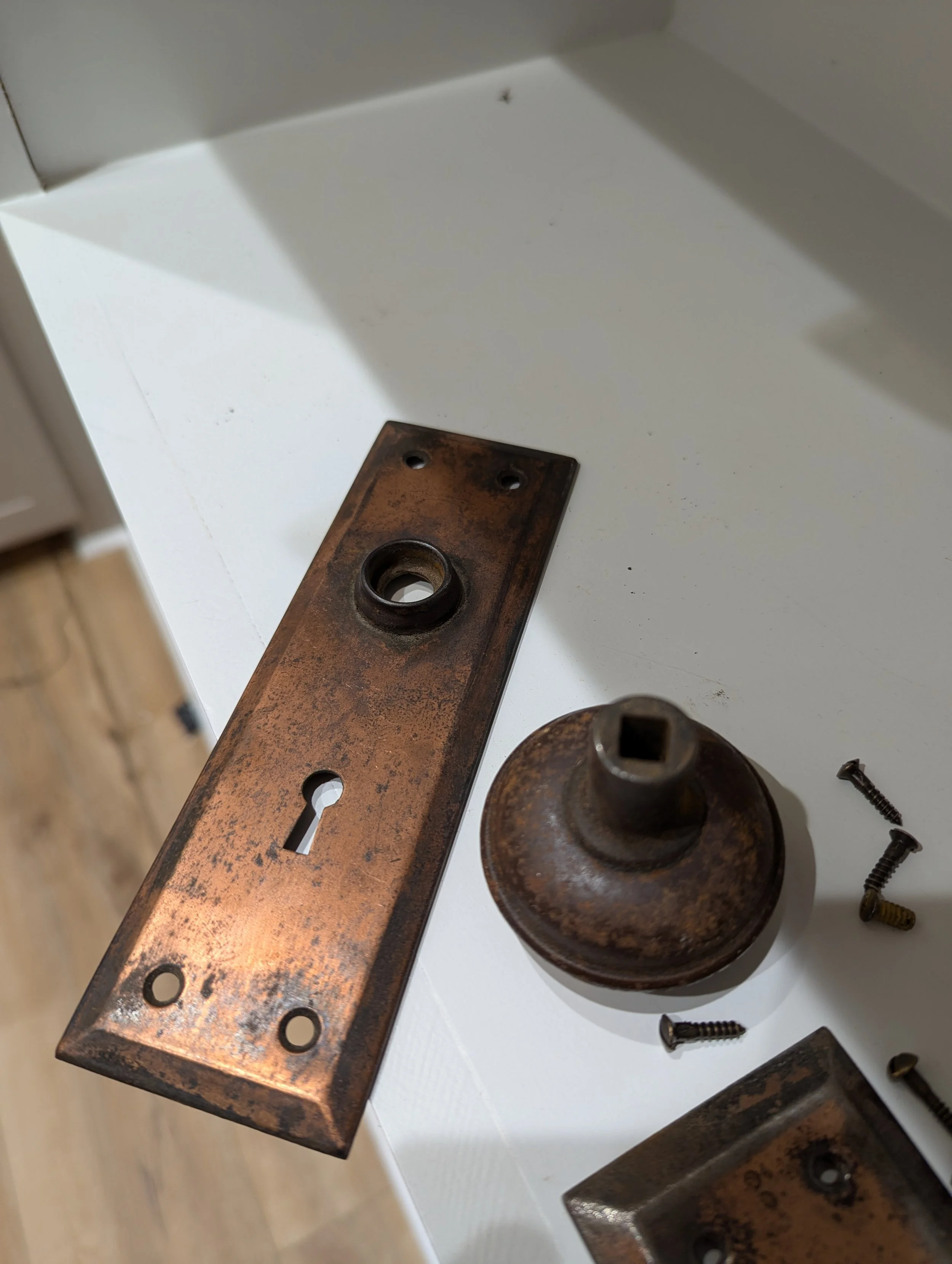 Disassembled antique door lock mechanism with a rusty lock plate, a round lock cylinder, and several screws on a white surface.