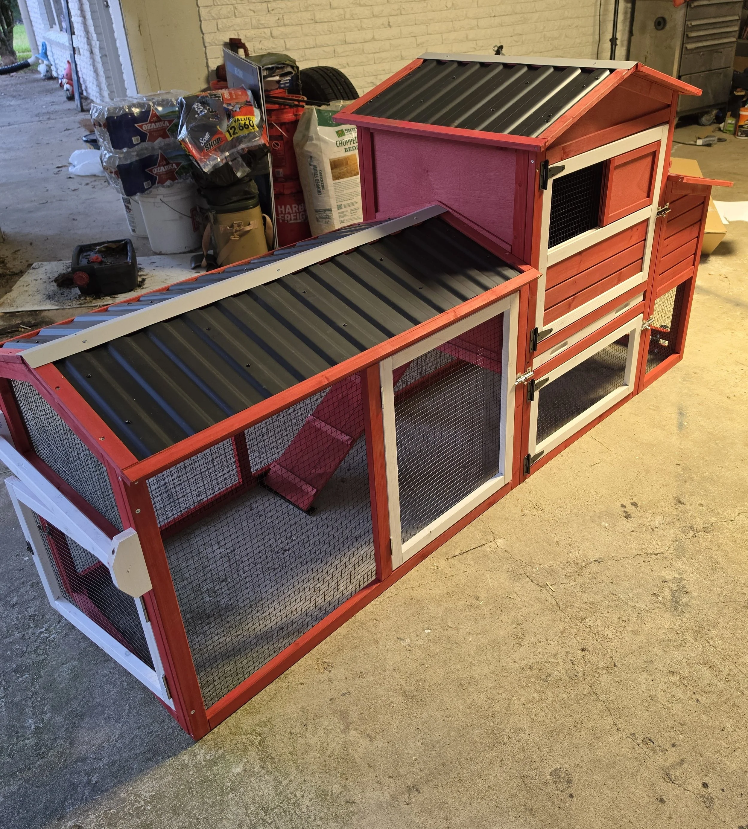 A red and white multi-level animal cage with a ramp, situated on a concrete floor in a garage or workshop. The cage has wire mesh sides and a pitched roof.