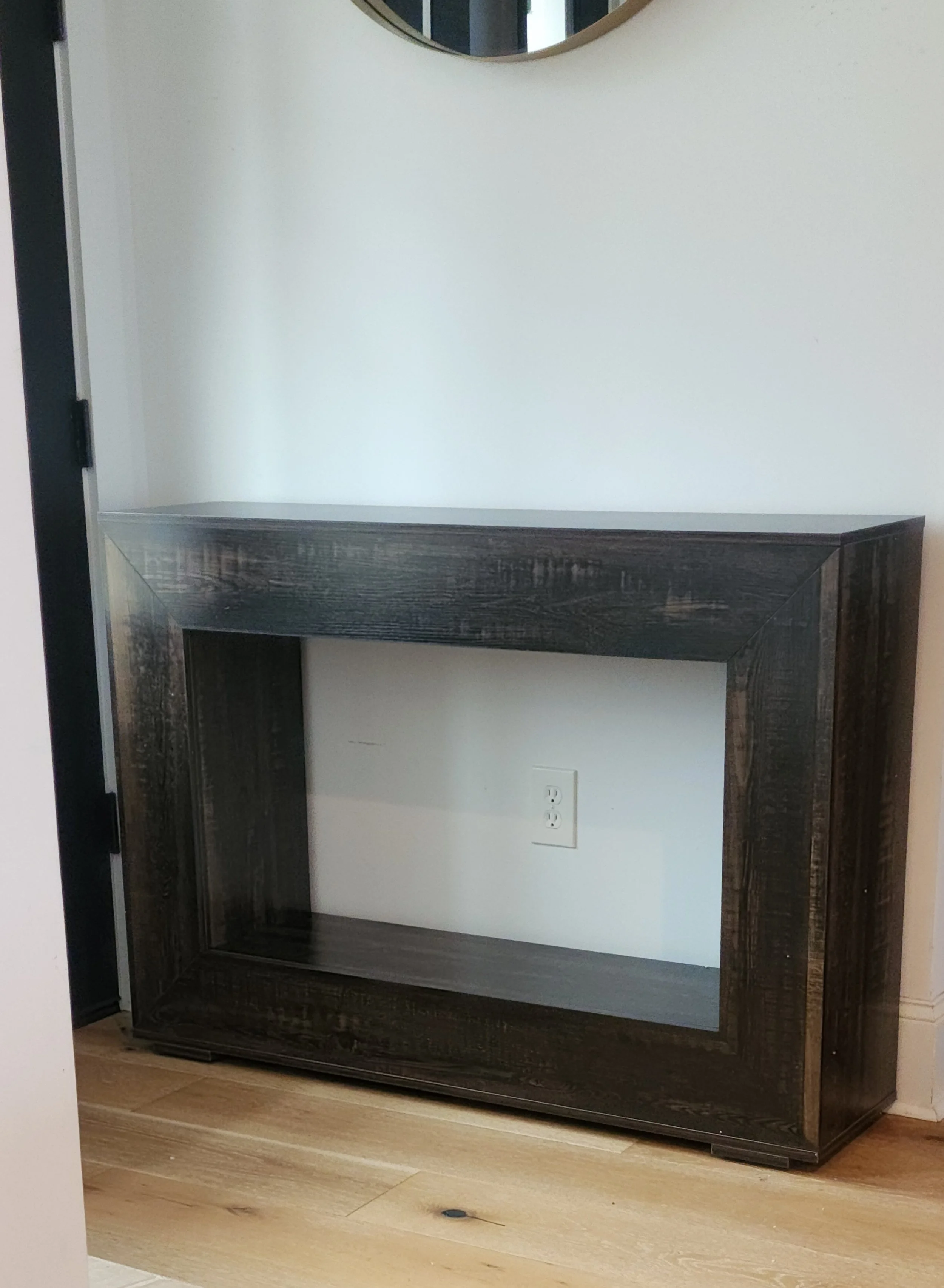 Empty black wooden shelf unit in a room with light-colored walls and wooden flooring, with an electrical outlet on the wall below the shelf.
