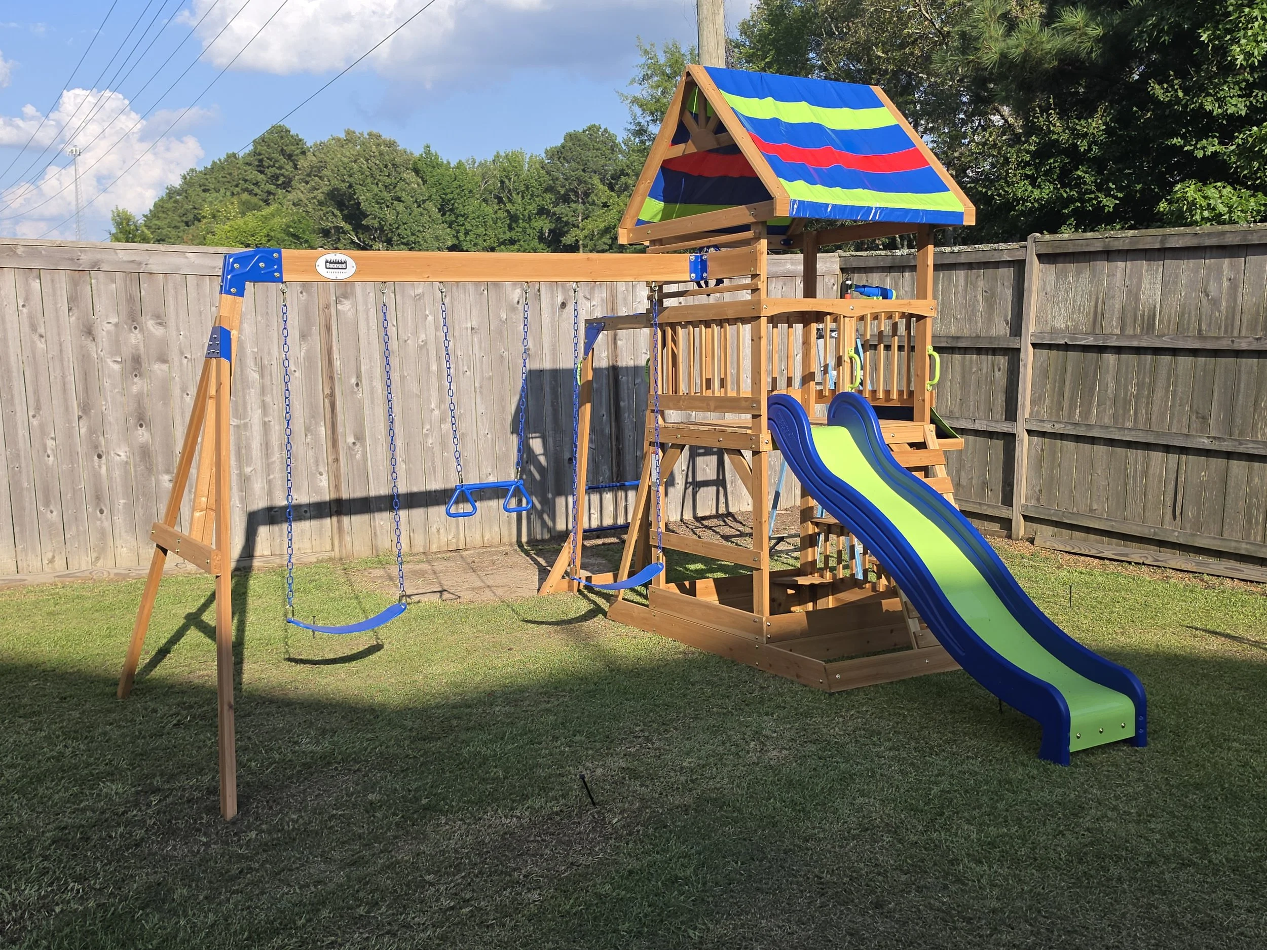 A backyard playground with a wooden swing set, two blue swings, a slide, and a playhouse with a colorful striped roof, enclosed by a wooden fence and surrounded by green trees.