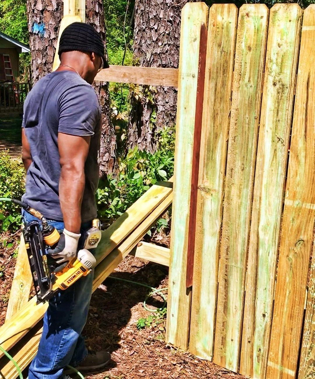 A man wearing a dark t-shirt, jeans, and a black cap is constructing a wooden fence using a nail gun during daytime. He is standing outdoors near trees.