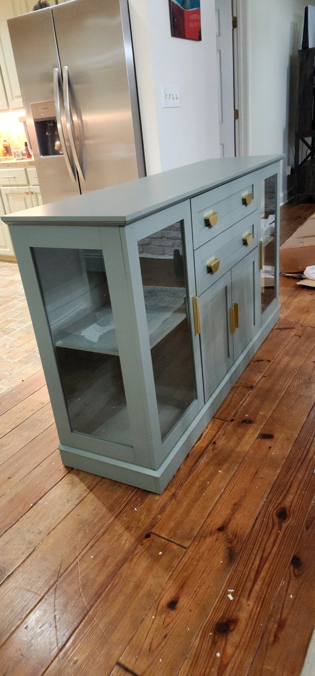 A small display cabinet with glass doors and drawers, painted light gray, with gold handles, placed on a wooden floor in a room.