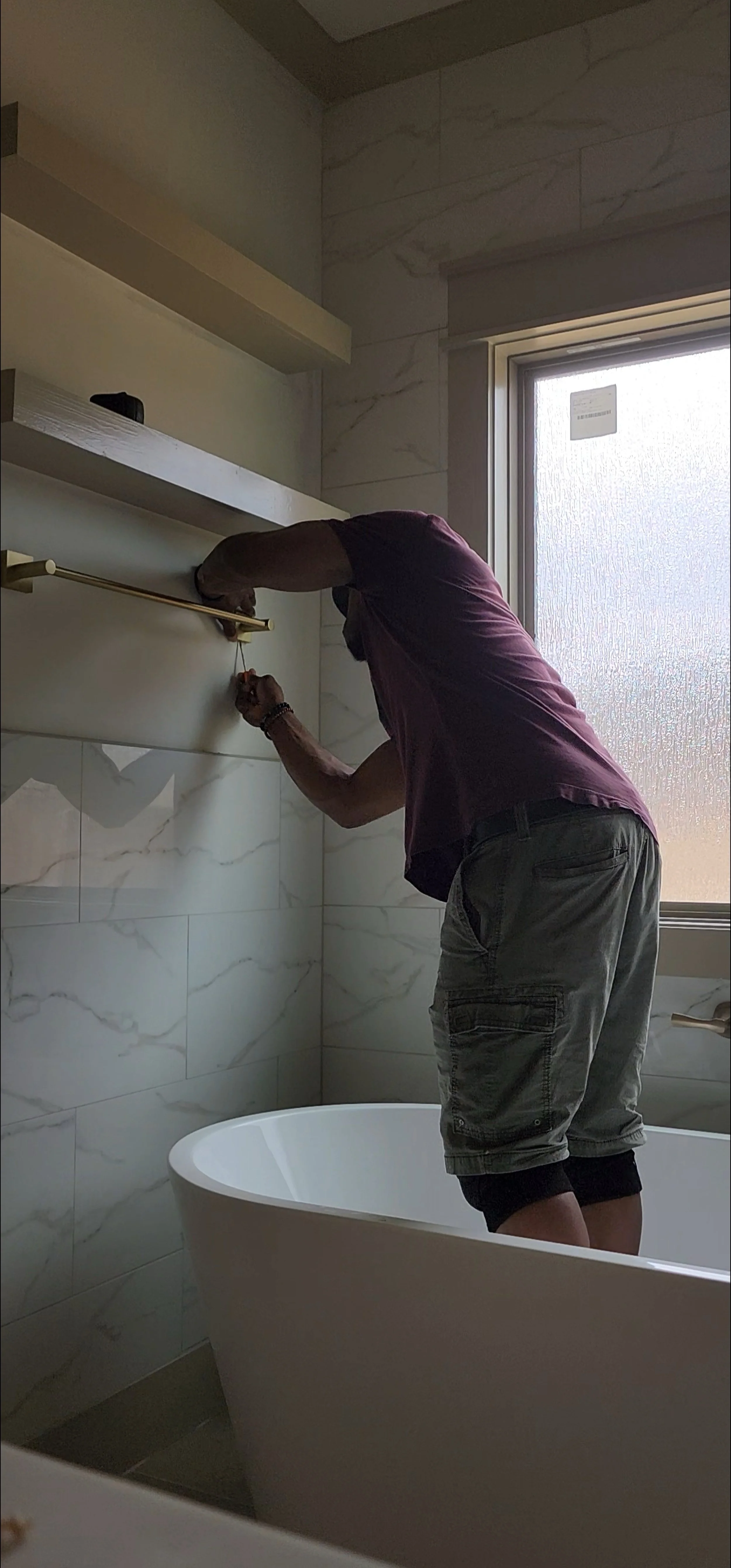 A man standing in a bathroom near a bathtub, reaching up to install or adjust a shelf on the wall, with a window nearby letting in natural light.