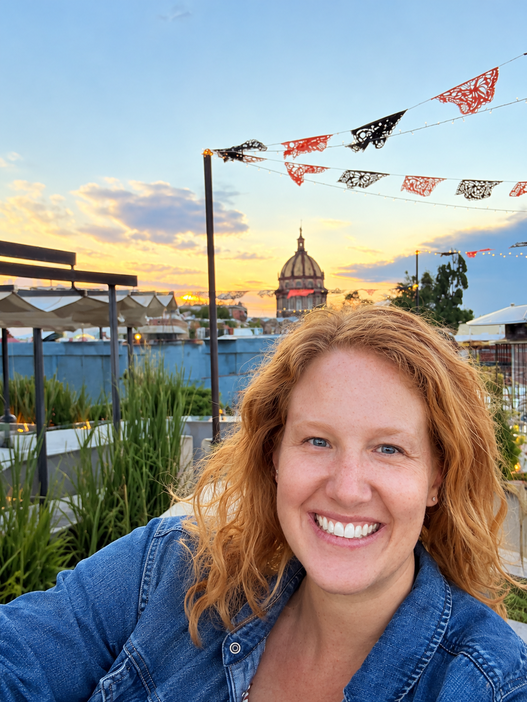 A woman with red curly hair taking a selfie outdoors during sunset with a cityscape and a large domed building in the background, decorative flags above, and plants around.