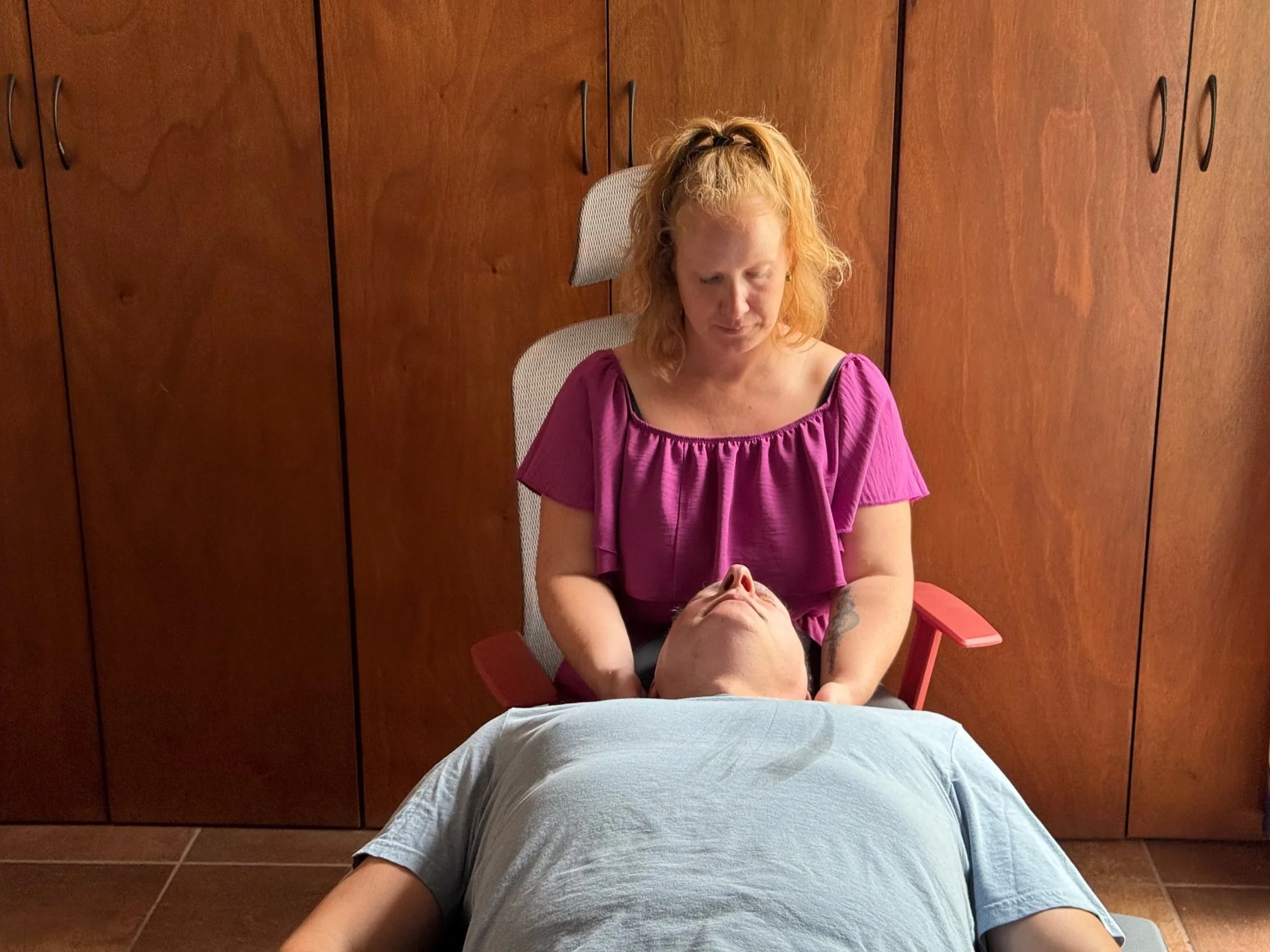 A woman with curly, red hair is sitting behind a person lying on a massage table. The massage table is covered with a white sheet. The woman is wearing a purple top and appears to be performing a massage. The room has wooden cabinets or closets in the background.