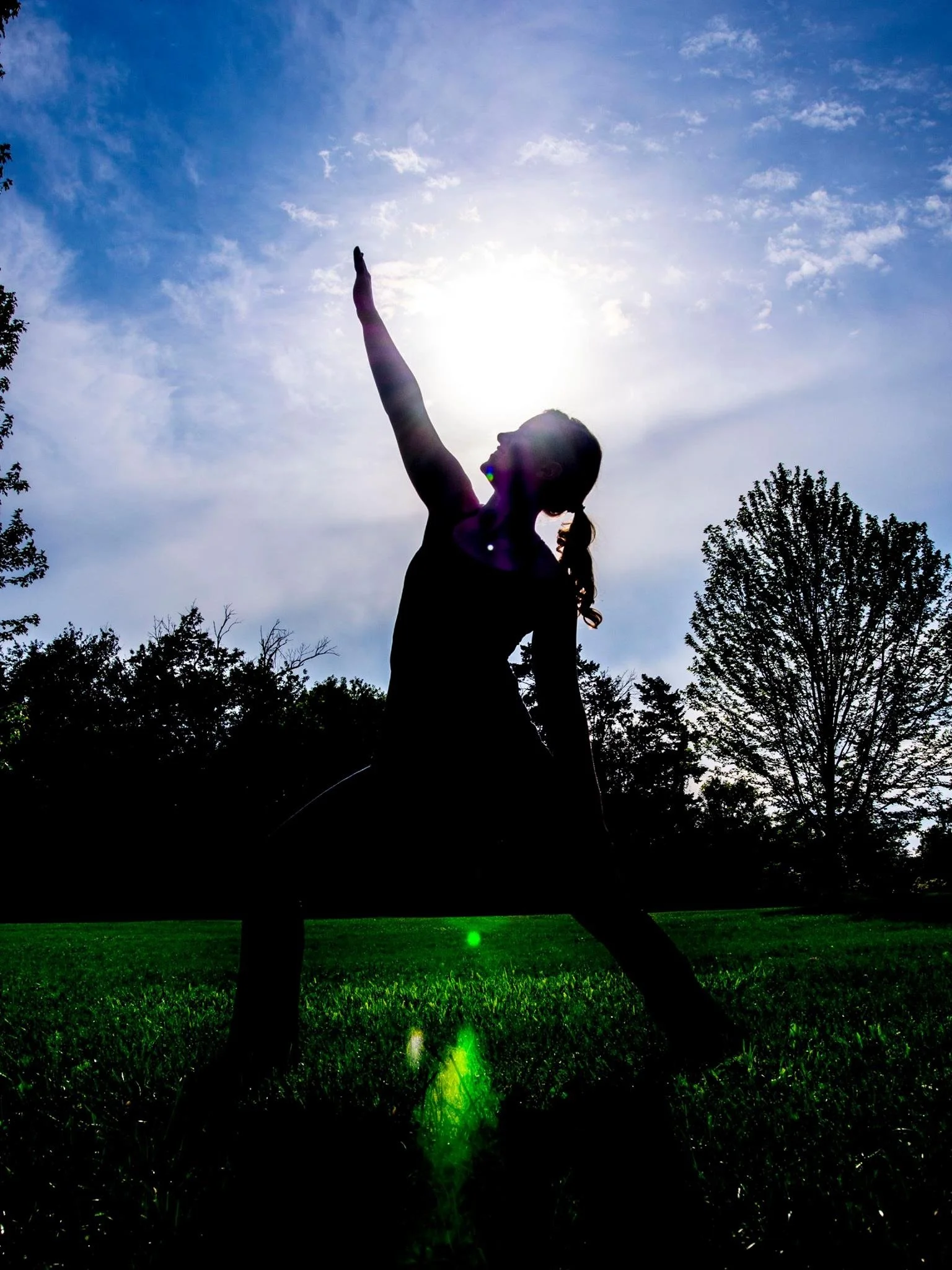 Silhouette of a woman doing yoga outdoors in a park during sunset, with green grass in the foreground and trees in the background.
