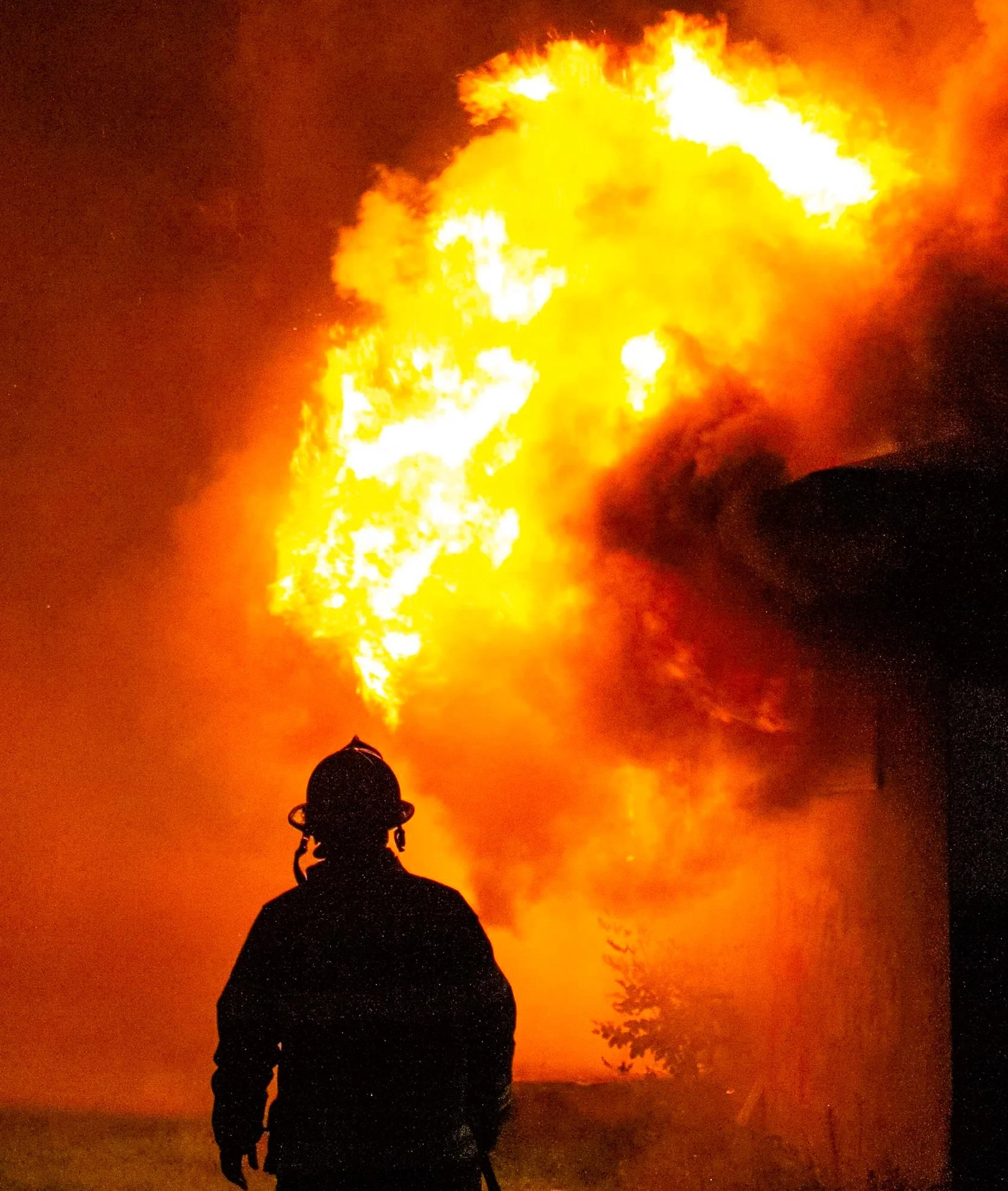 Silhouette of a person wearing a hat, standing in front of a large fire or explosion with bright orange and yellow flames illuminating the night sky and some trees in the background.