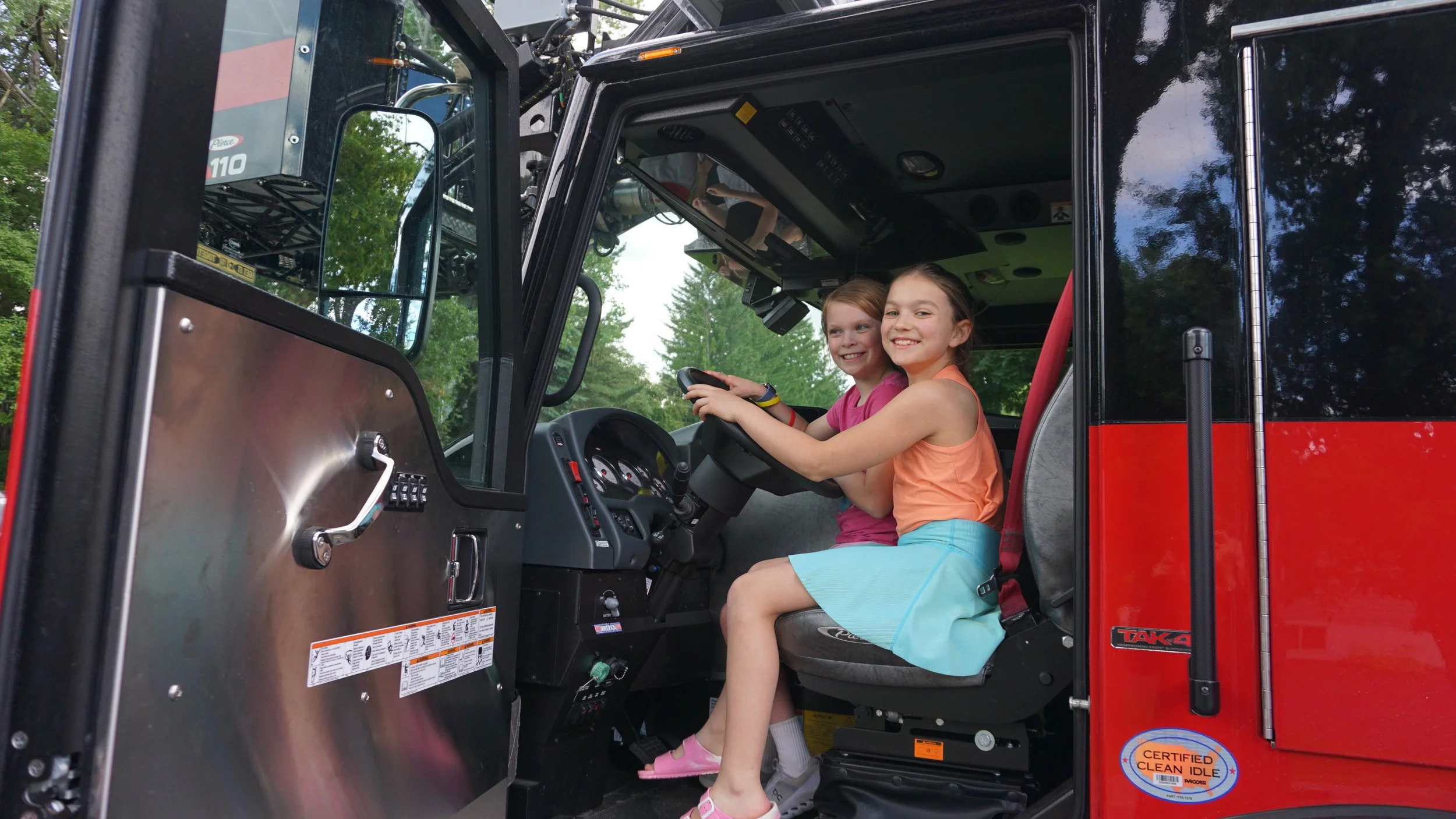 Two young girls sitting in the cab of a fire truck, smiling. One girl has reddish hair and the other has dark brown hair. They are surrounded by lush green trees.