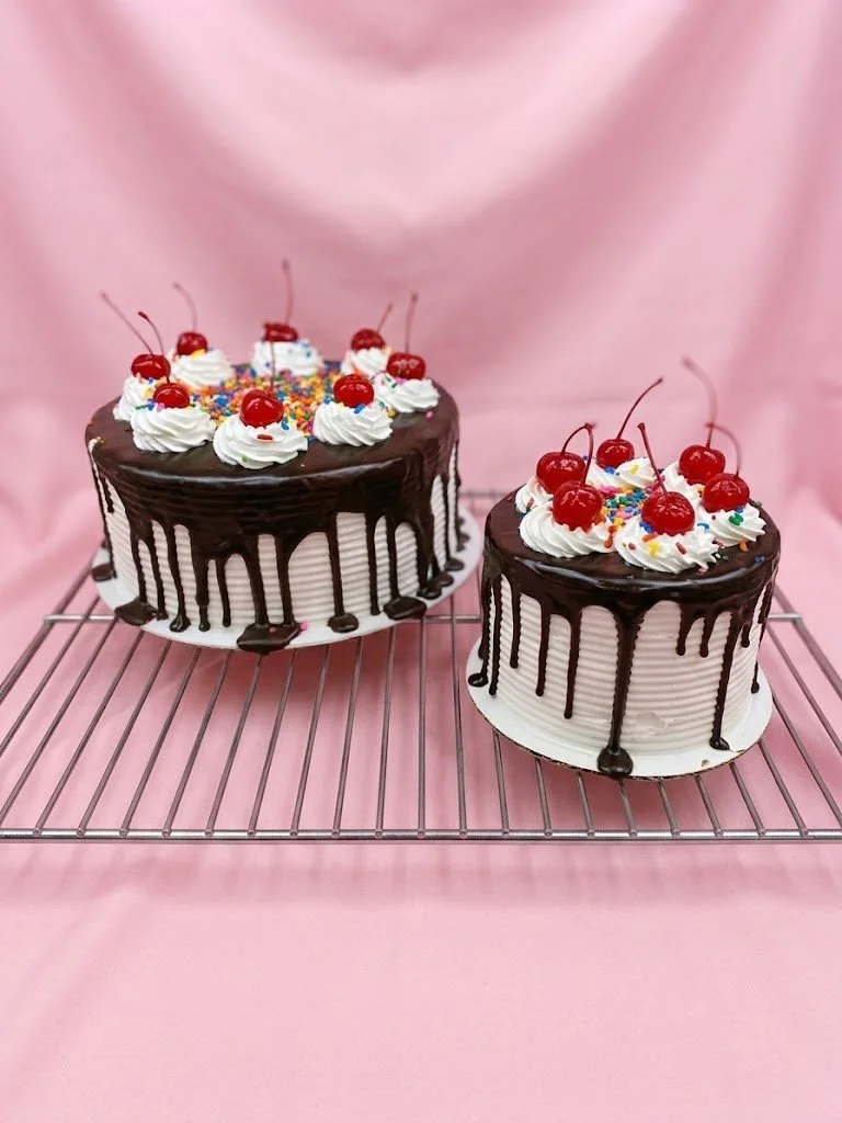 Two decorated cakes with chocolate drizzle, whipped cream, cherries, and sprinkles on a cooling rack against a pink background.