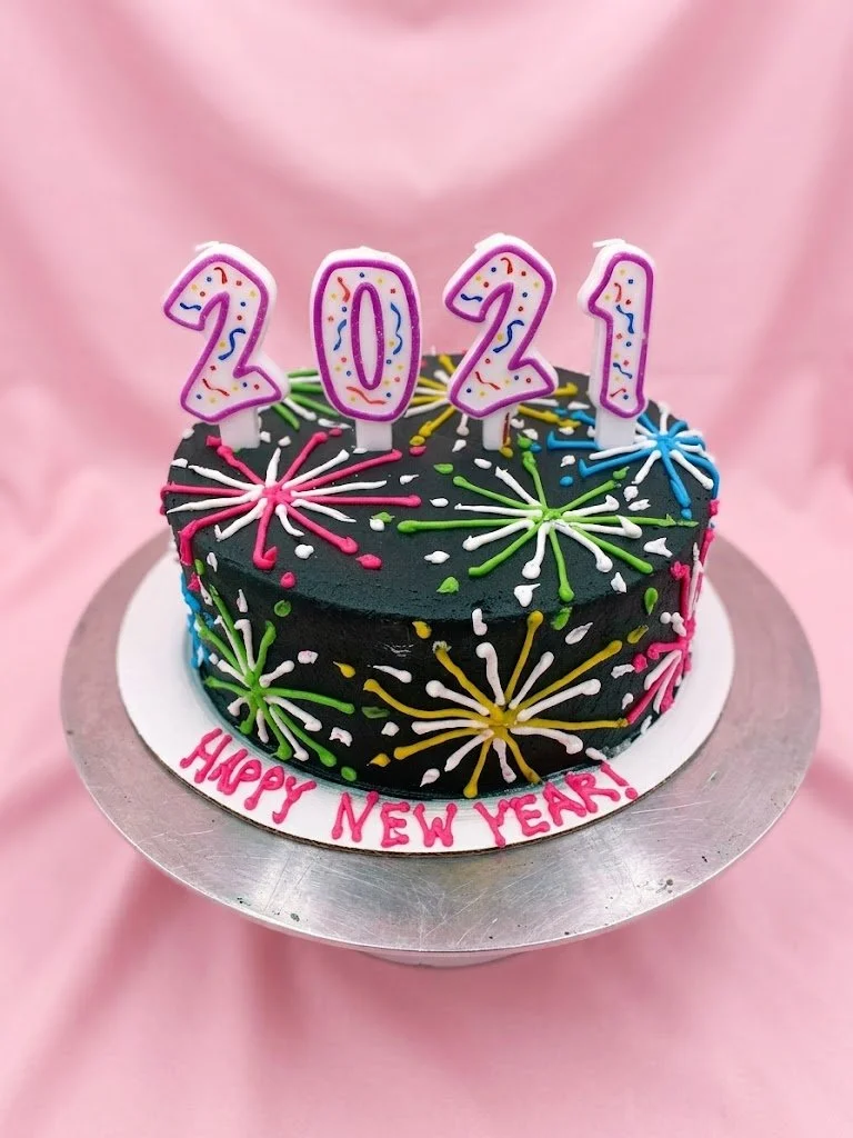 Chocolate cake decorated with colorful sprinkles and fireworks designs, with four pink candles spelling out 2021, and a message that reads "Happy New Year!"