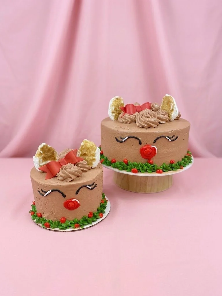 Two reindeer-themed cakes decorated with chocolate frosting, red and green icing, and candy decorations, placed on a pink background.