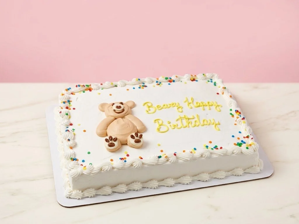 Rectangular white birthday cake with colorful sprinkles and a teddy bear decoration on top, inscribed with "Beary Happy Big Birthday" in yellow icing, on a white surface against a pink background.