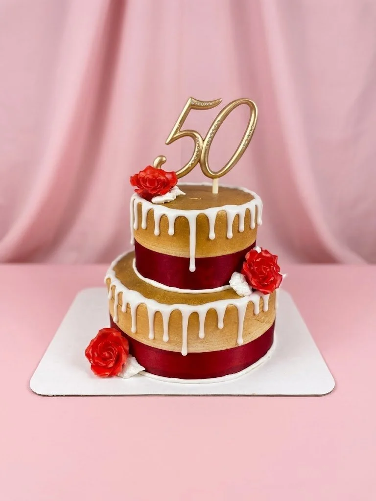 A two-tiered birthday cake with white drip icing, red ribbon sides, and red roses on top and sides, decorated for a 50th celebration with a gold "50" topper against a pink background.