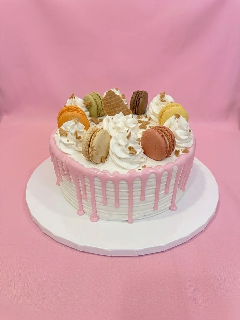 A round cake decorated with white frosting, pink icing drips, and macarons in various pastel colors on top, along with whipped cream swirls and small pieces of chopped nuts or chocolate.