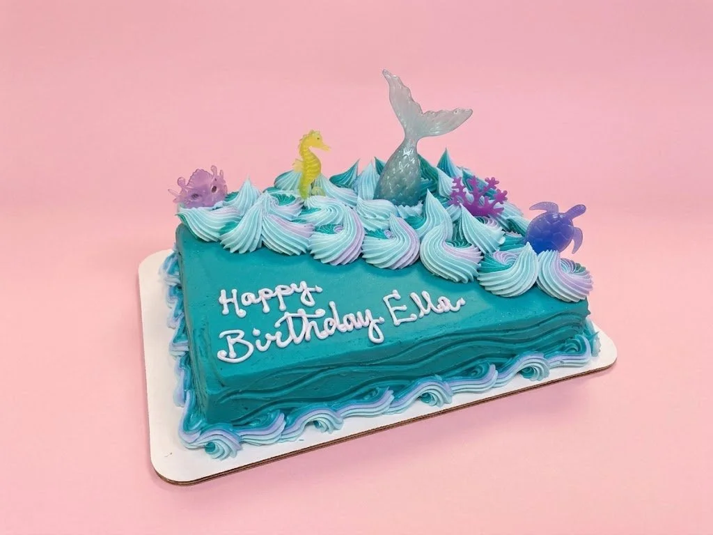 A birthday cake with an ocean and mermaid theme, decorated with sea creature toppers, blue and purple swirls, and a mermaid tail on top, with a message that says "Happy Birthday Ella".