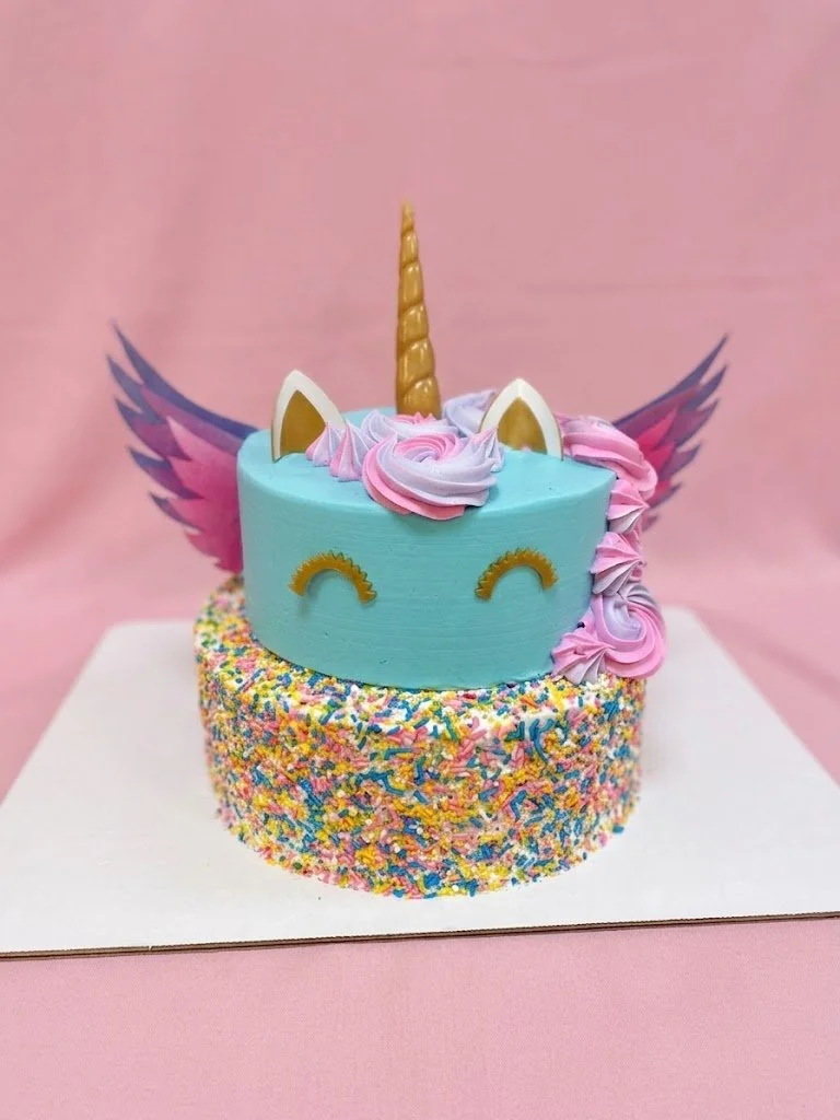 Colorful unicorn-themed birthday cake with a blue unicorn face, golden horn, pink and purple swirled frosting mane, white ears with gold accents, pink and purple wings, and multicolored sprinkles around the base, against a pink background.