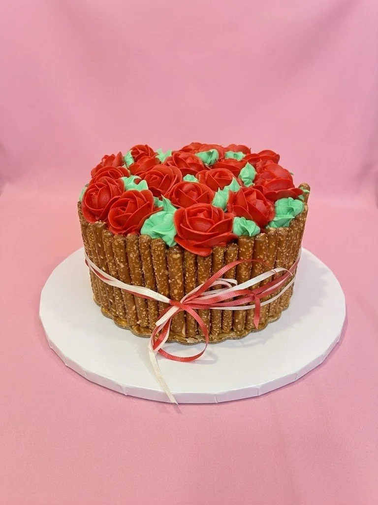 Round cake decorated with red and green frosting, surrounded by stacked pretzels, tied with pink and cream ribbons.