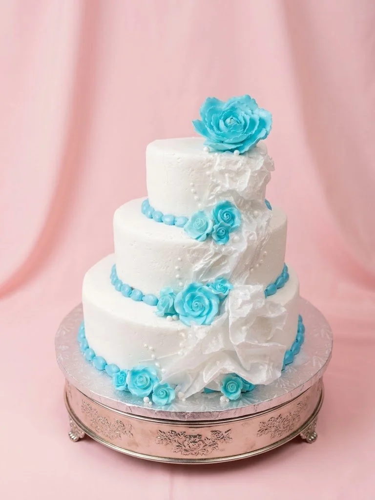 Three-tier white wedding cake decorated with blue sugar roses and a large blue flower on top, placed on a silver cake stand with floral engravings.