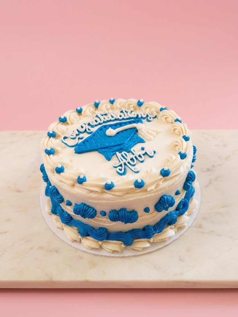 A round graduation cake with white and blue decorations, featuring a graduation cap and a diploma on top, with the words 'Congratulations Aori' written in blue icing.