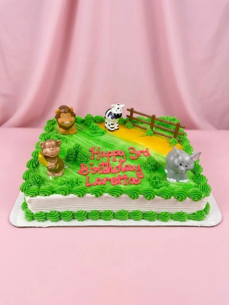 Animal-themed birthday cake with green snake-like frosting, animal figurines including a lion, zebra, monkey, and elephant, and a pink message saying 'Happy 3rd Birthday Loretta' on top, set against a pink curtain background.