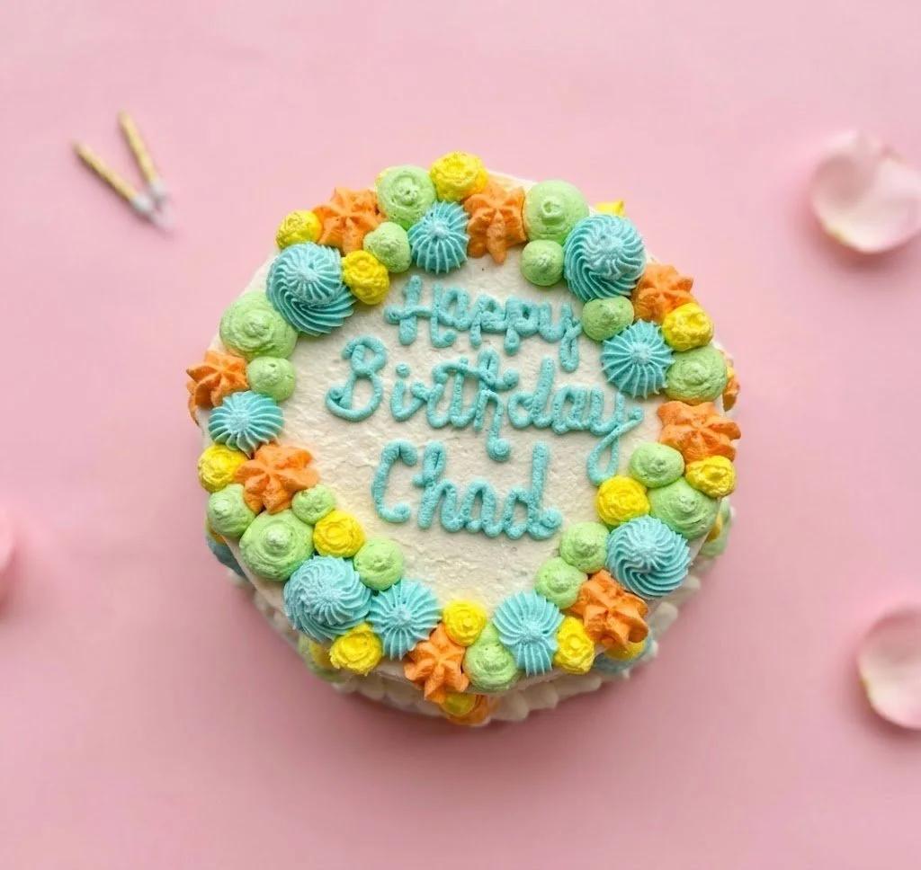 A birthday cake with colorful icing decorations in blue, yellow, orange, and green, with the message "Happy Birthday Chad" written in blue icing on top, placed on a pink background with some flower petals around it.
