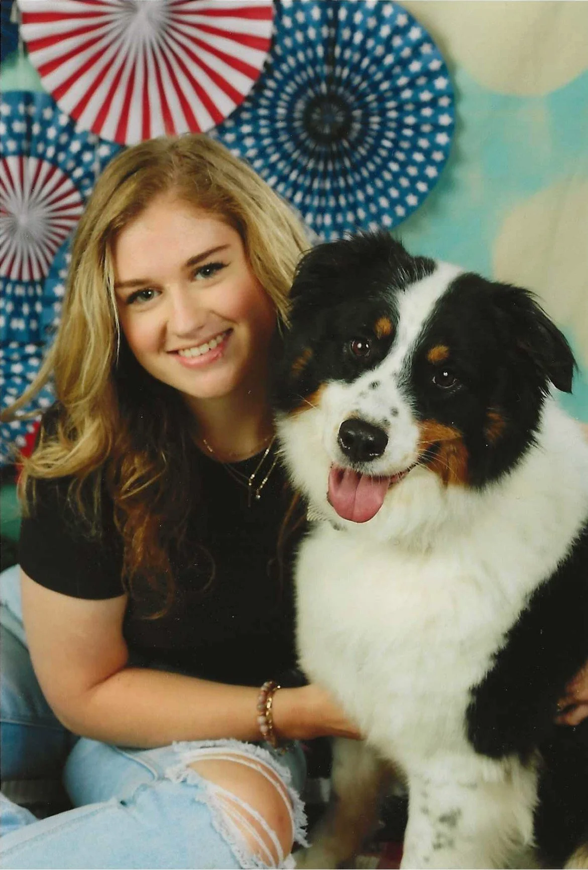 A young woman with long blonde wavy hair smiling and sitting next to a large Bernese Mountain Dog with its tongue out. The background features patriotic-themed parasols in red, white, and blue.