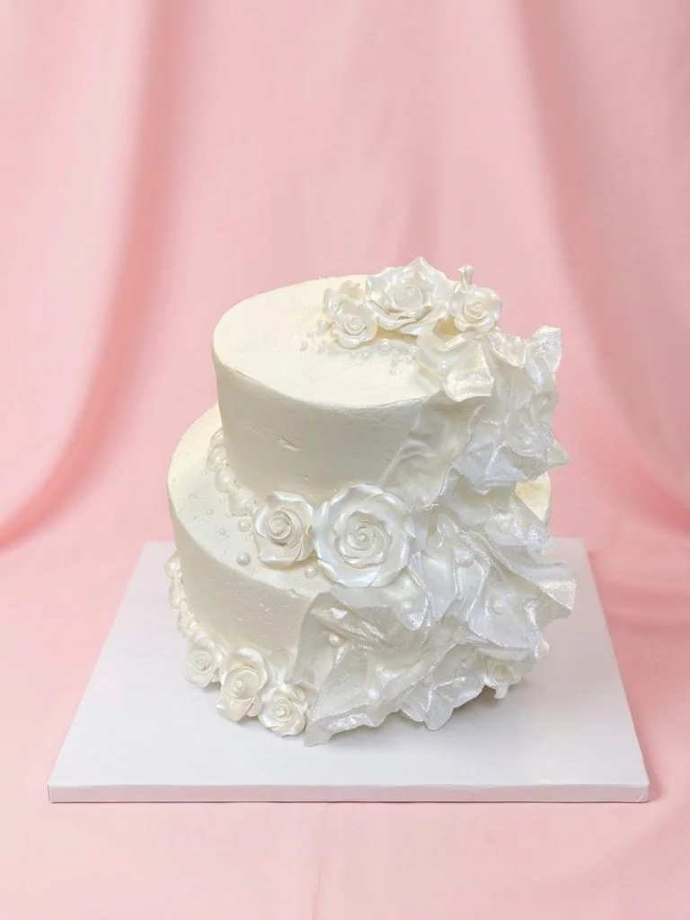 A white, two-tier wedding cake decorated with white roses and large white ruffles, set against a pink backdrop.