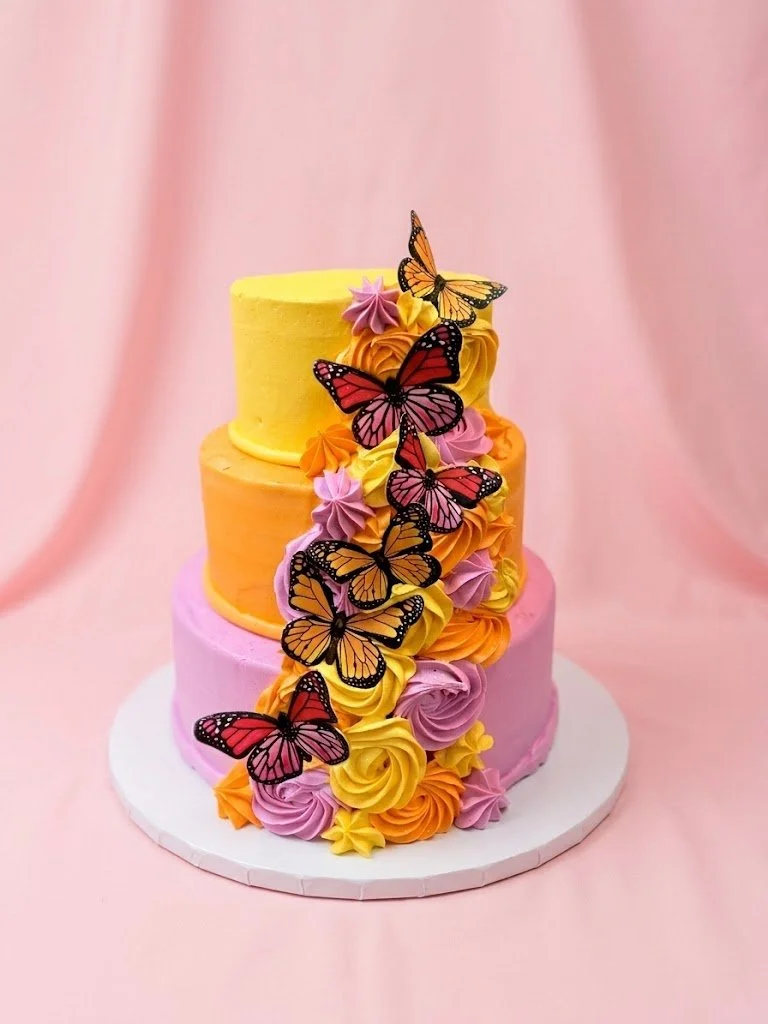 Three-tiered pastel-colored cake with yellow, orange, and pink icing, decorated with pink, purple, yellow, and orange whipped frosting rosettes, and topped with edible butterfly decorations, all set against a soft pink background.