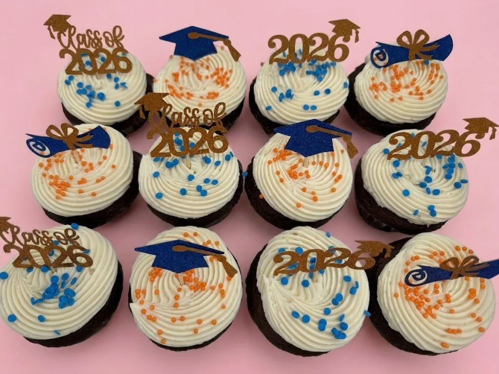 A display of cupcakes decorated with white frosting, orange and blue sprinkles, and toppers that say 'Class of 2026,' featuring blue graduation caps and gold '2026' signs, arranged on a pink surface.